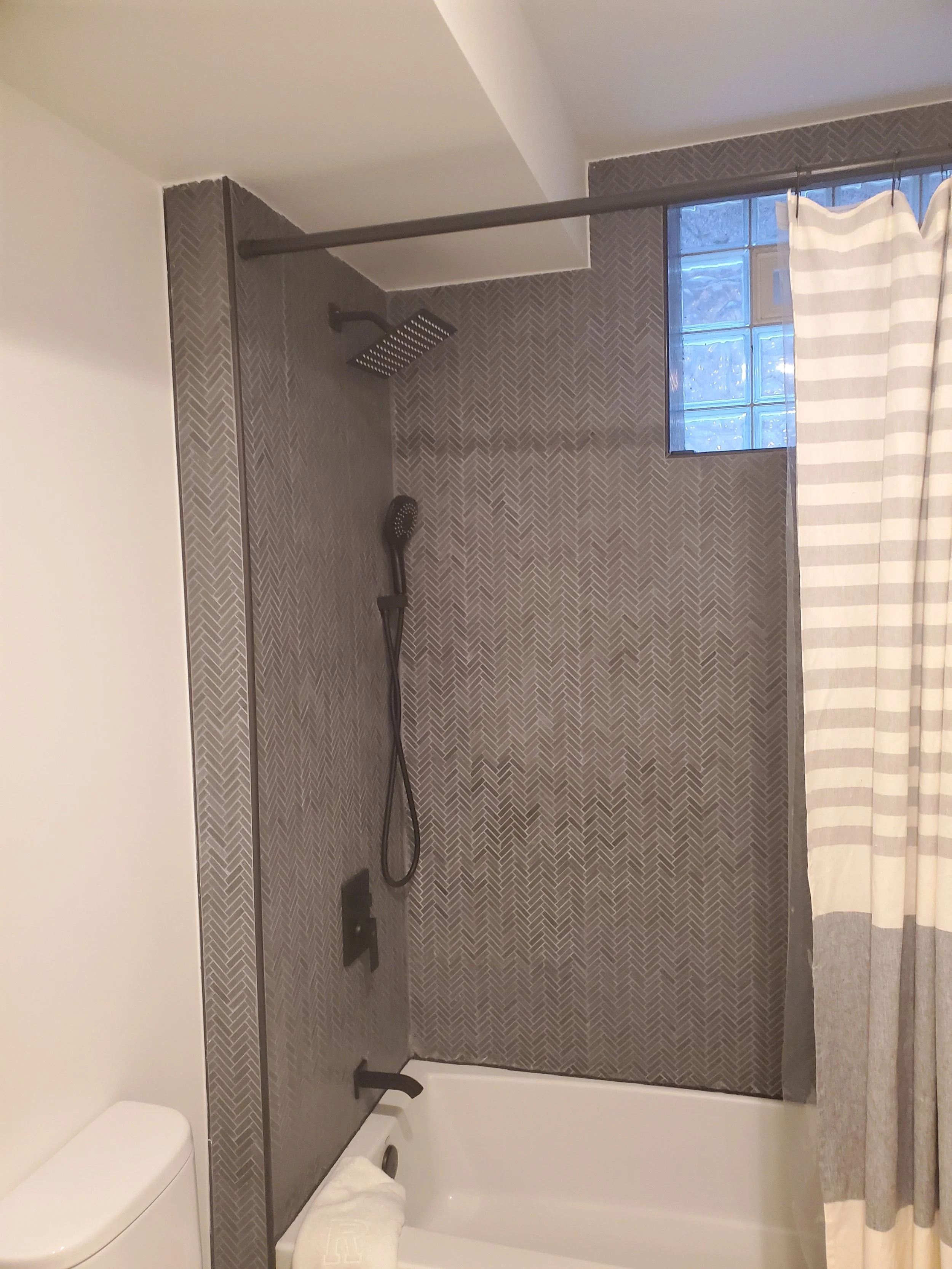 A modern bathroom with a walk-in shower featuring a dark herringbone tile wall, a black showerhead, a handheld shower, and a glass block window. A black shower rod has a hung striped white and beige curtain.