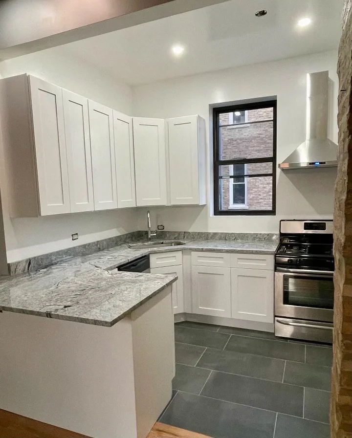 Modern kitchen with white cabinets, granite countertops, black window, stainless steel oven, and gray tiled floor.