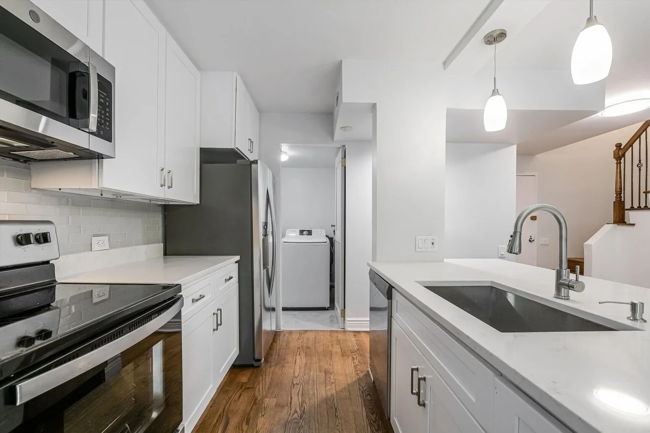 White kitchen with stainless steel appliances, including microwave and refrigerator, and a laundry area with a washing machine visible through an open door. Wooden flooring and modern pendant lighting above a white countertop with a black sink.