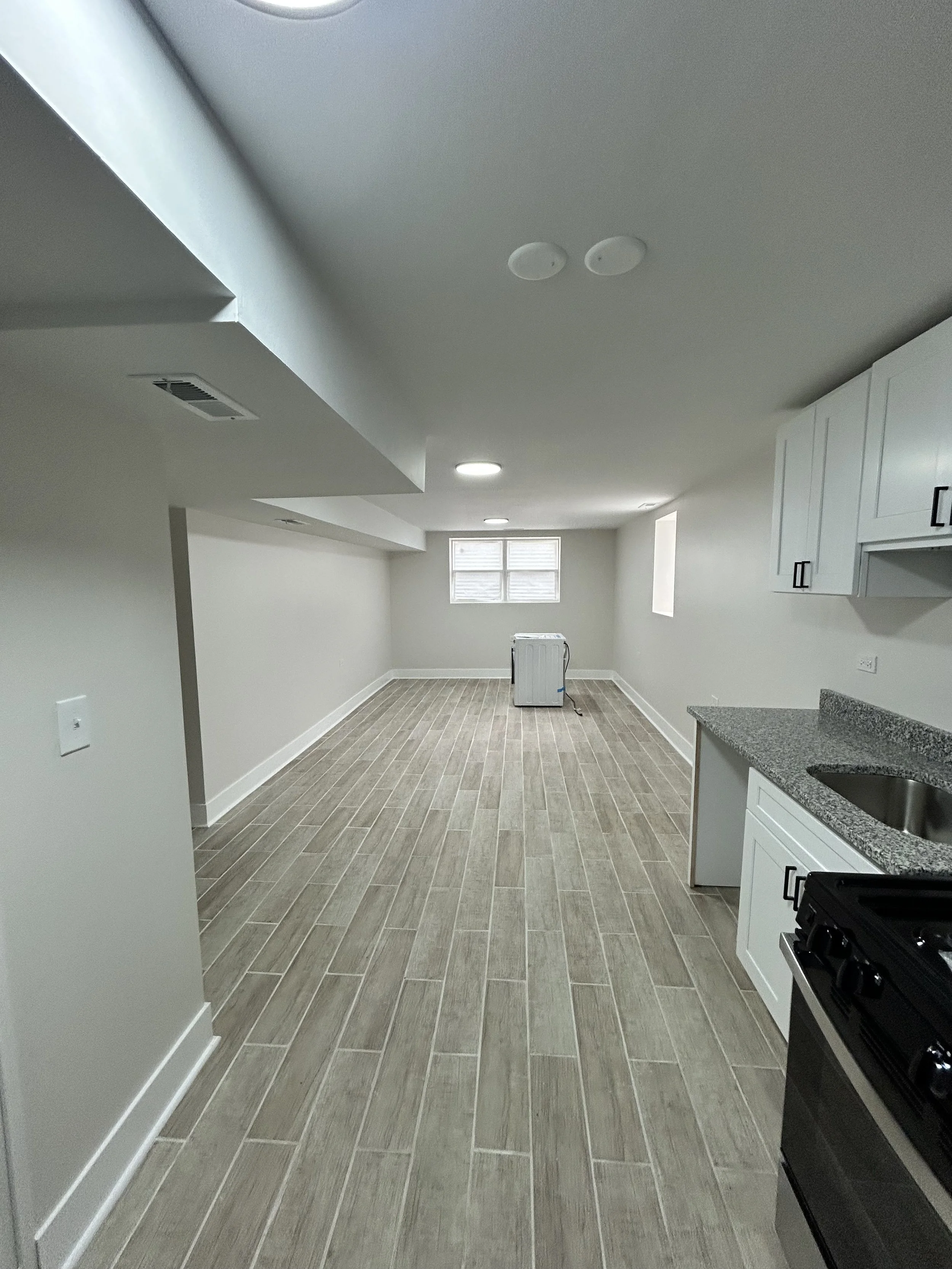 Empty kitchen with gray wood-look tile flooring, white cabinets with black handles, a black stove, granite countertops, a small window, and some appliances in the background.