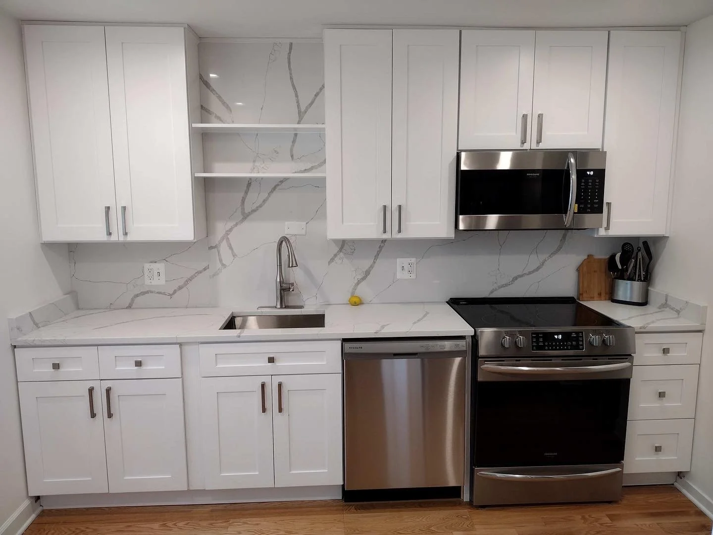 Modern white kitchen with marble backsplash and countertops, stainless steel appliances including a microwave, dishwasher, and oven, white upper and lower cabinets, and a wooden cutting board with utensils on the counter.