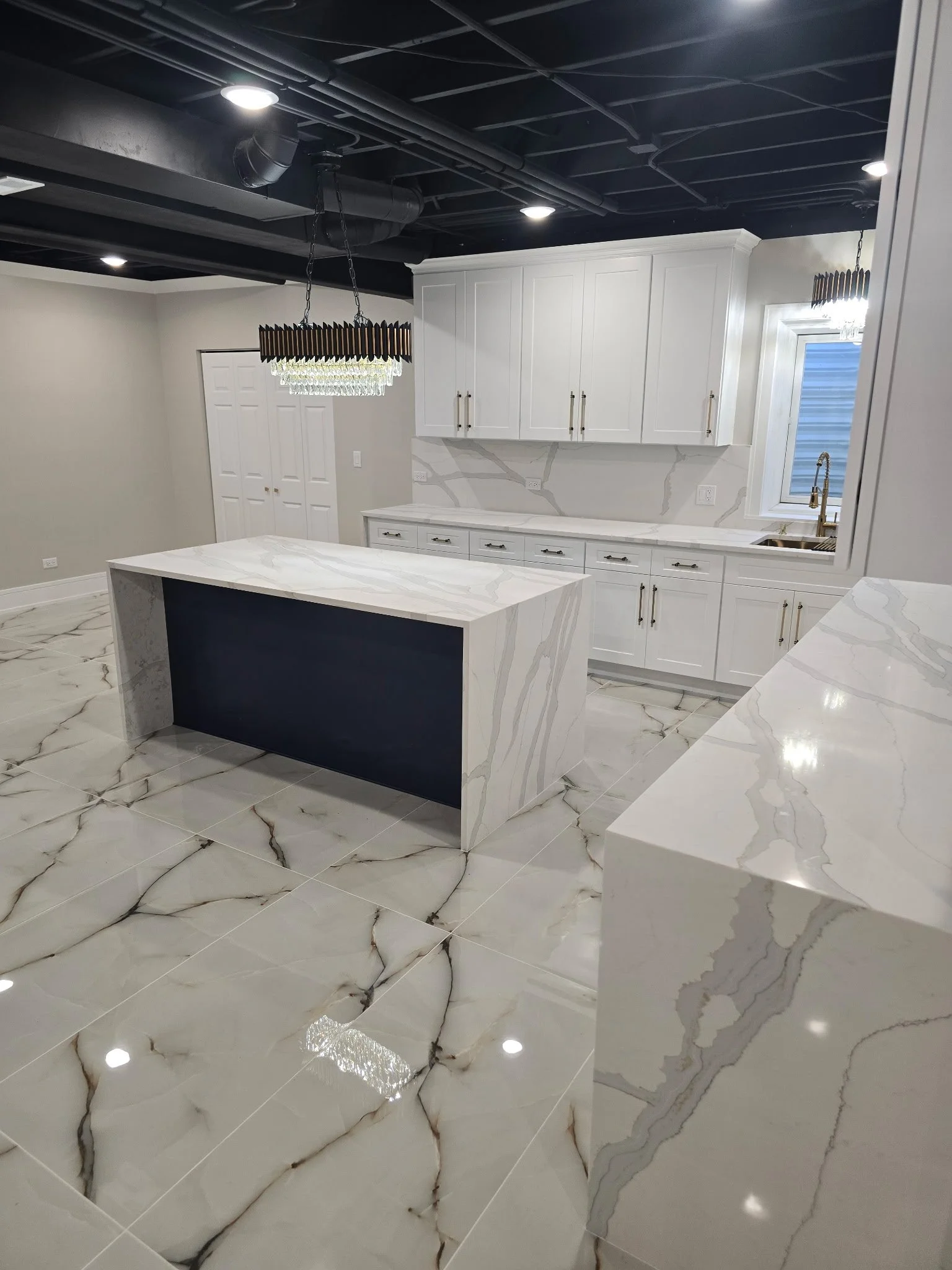 Modern kitchen with white cabinetry, marble countertops, and large marble floor tiles; black ceiling with recessed lighting and two chandeliers; window near sink.