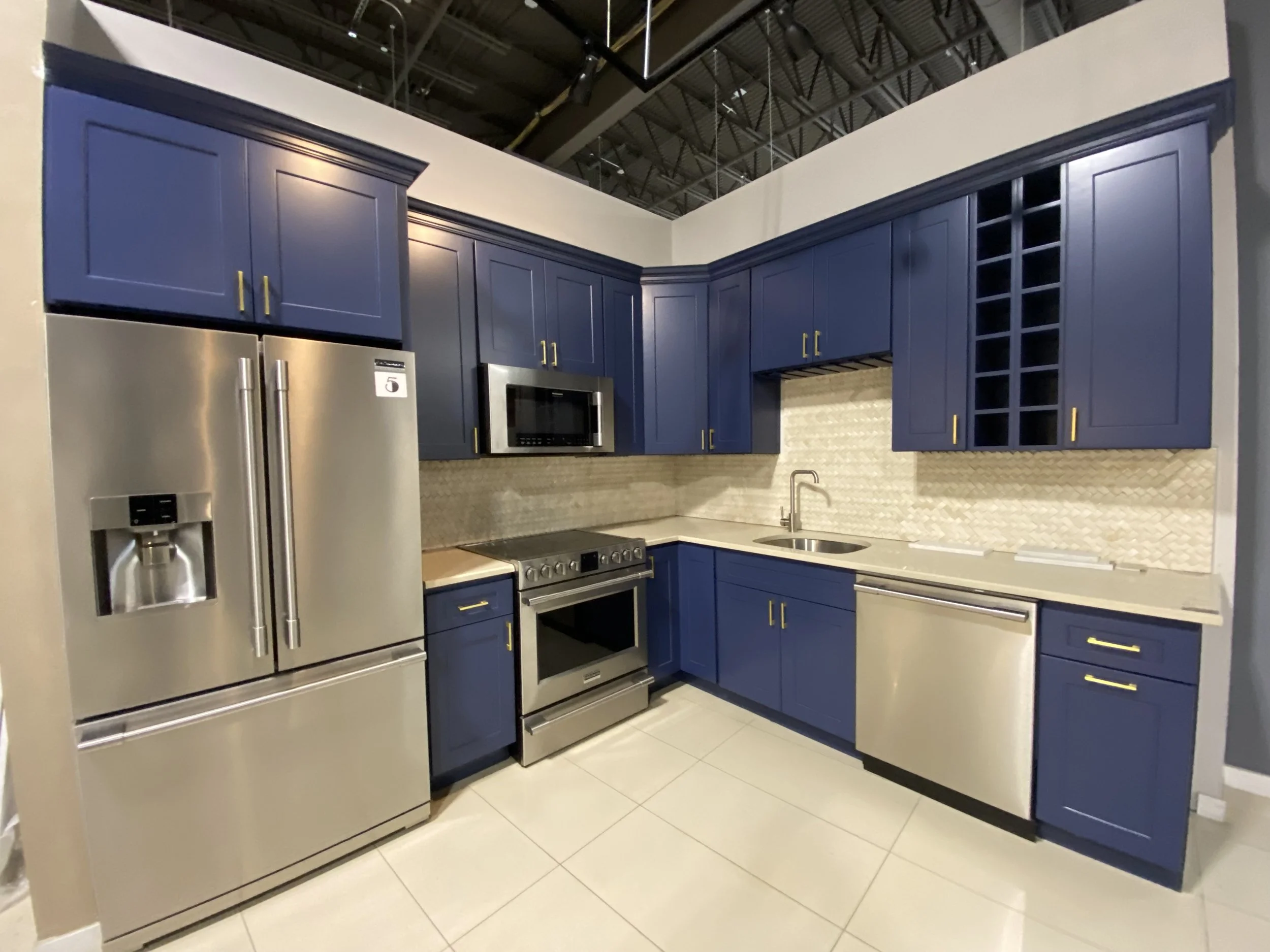 A kitchen display with blue cabinets, stainless steel refrigerator, microwave, and oven. The countertop has a sink, and there is a decorative tile backsplash. The ceiling is open with visible beams and ductwork.