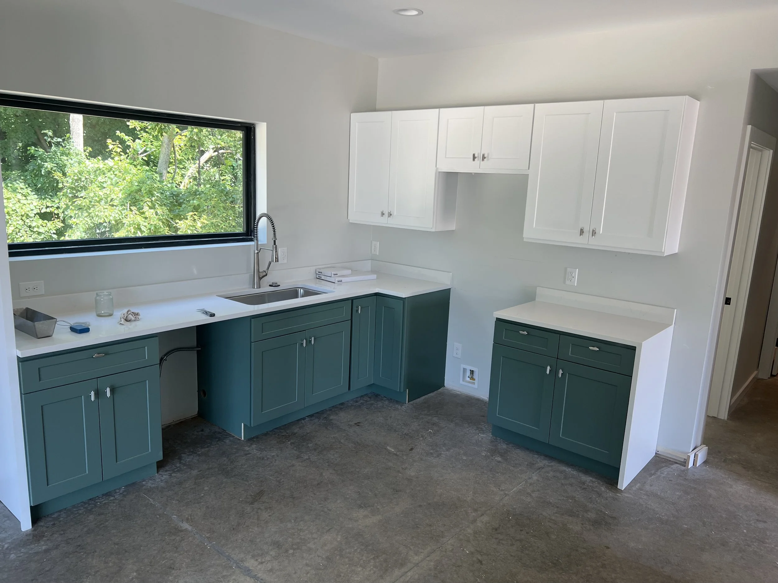 Unfinished kitchen with white upper cabinets and teal lower cabinets, a large window overlooking green trees, and an exposed concrete floor.