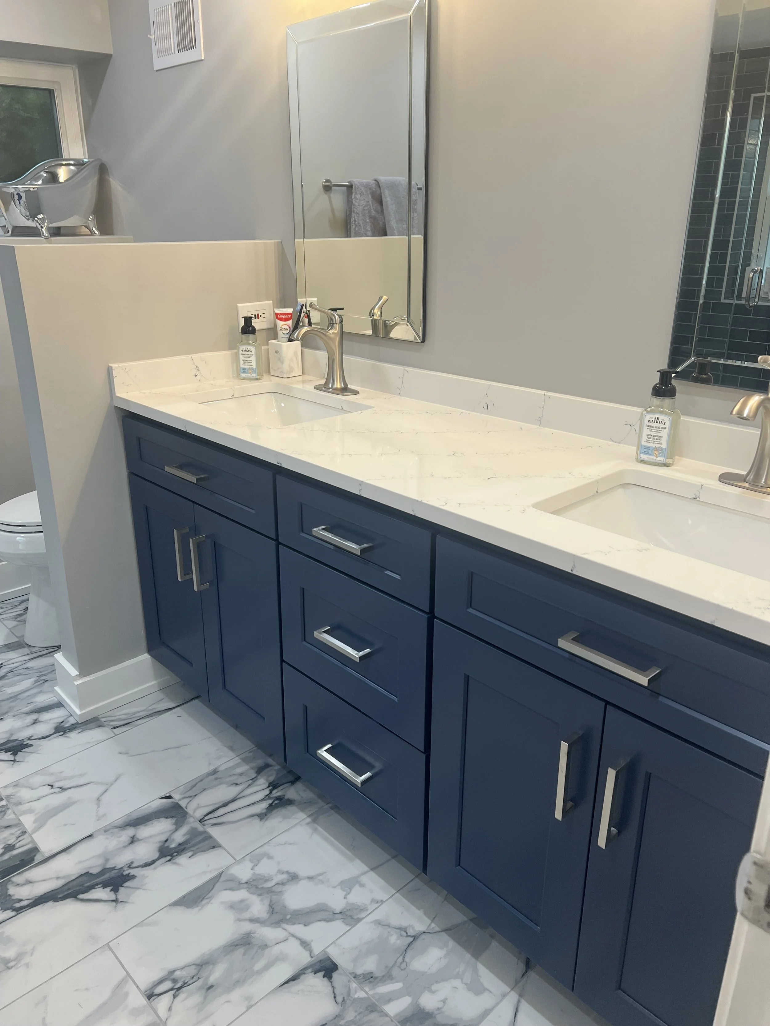 Bathroom with blue cabinetry, white marble countertop, a large mirror, and a marble tiled floor. Visible are two sinks, a soap dispenser, and a hand towel hanging on a rod. There is a window in the background, and a small silver decorative bathtub is