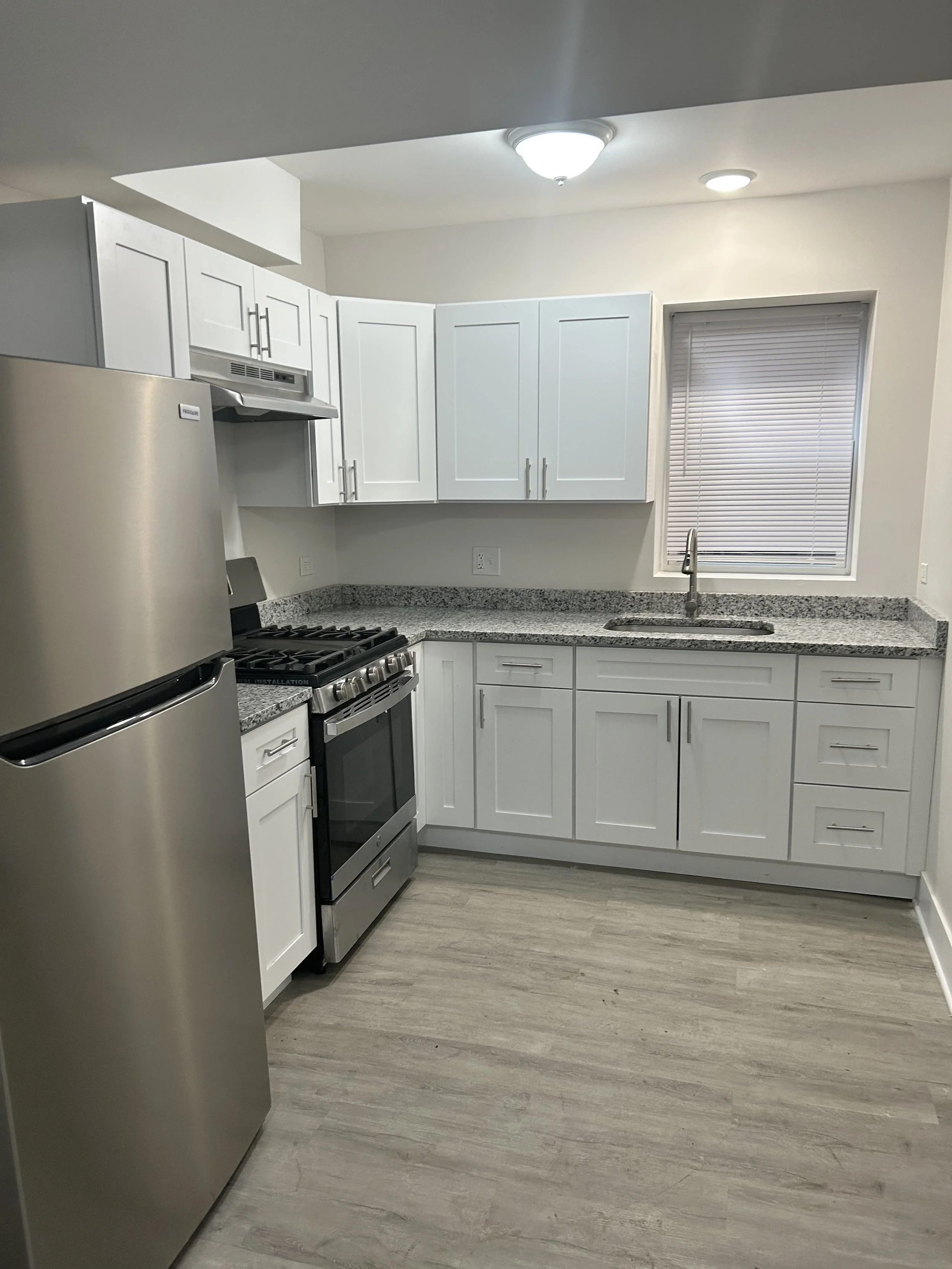A modern kitchen with white cabinets, stainless steel refrigerator and stove, granite countertops, a sink under a window with closed blinds, and wood-look flooring.