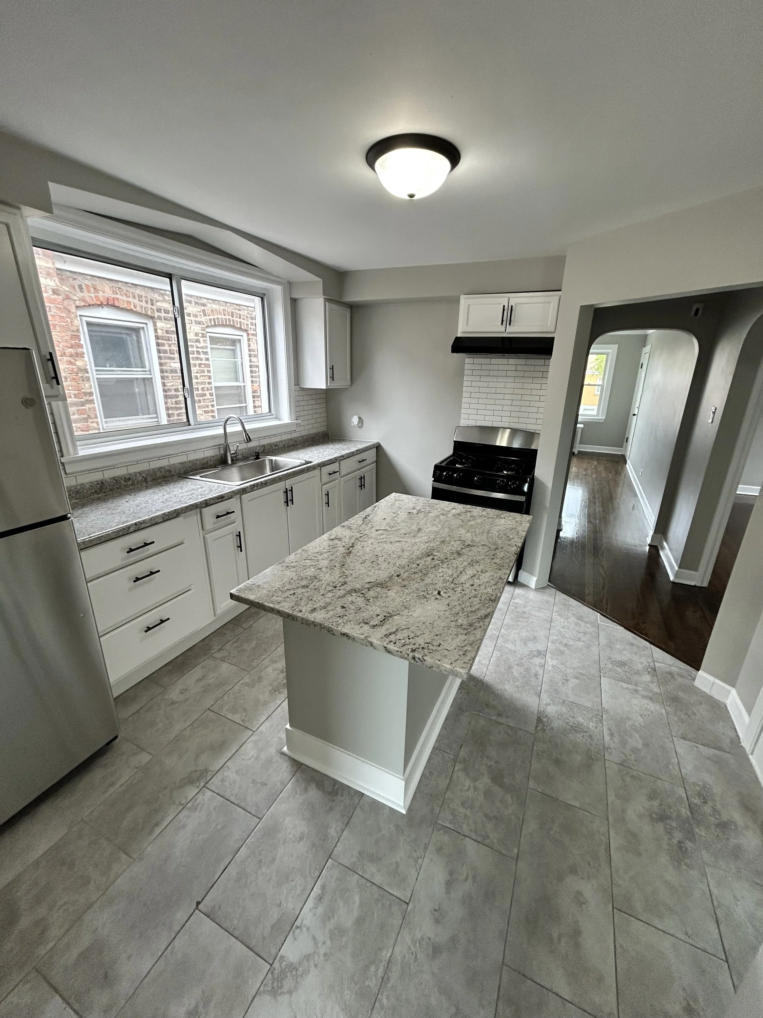 Kitchen with granite countertops, white cabinets, stainless steel appliances, tile flooring, large window, and a hallway leading to other rooms.