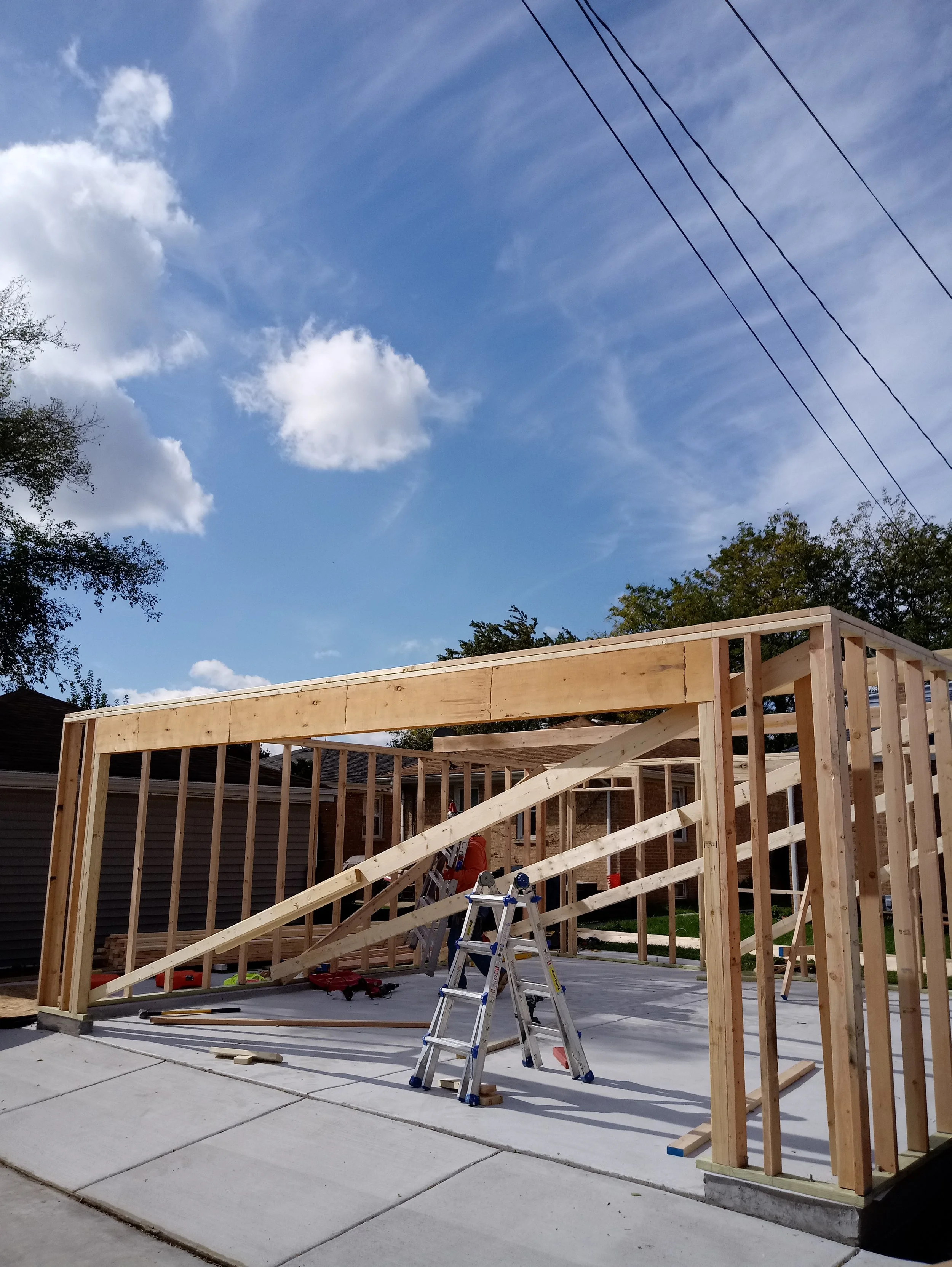 Wooden framing for a house under construction, with a partly assembled roof structure, ladder, and construction tools on a concrete patio, under a partly cloudy sky with trees and power lines in the background.