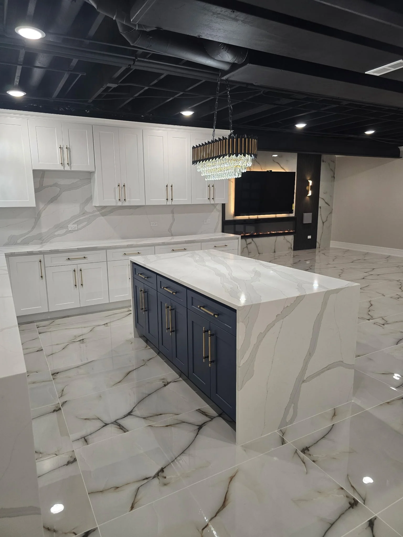 Modern kitchen with white cabinets, a blue island, marble countertops, a marble backsplash, a chandelier, and marble flooring, with a black ceiling featuring recessed lights and visible ductwork.