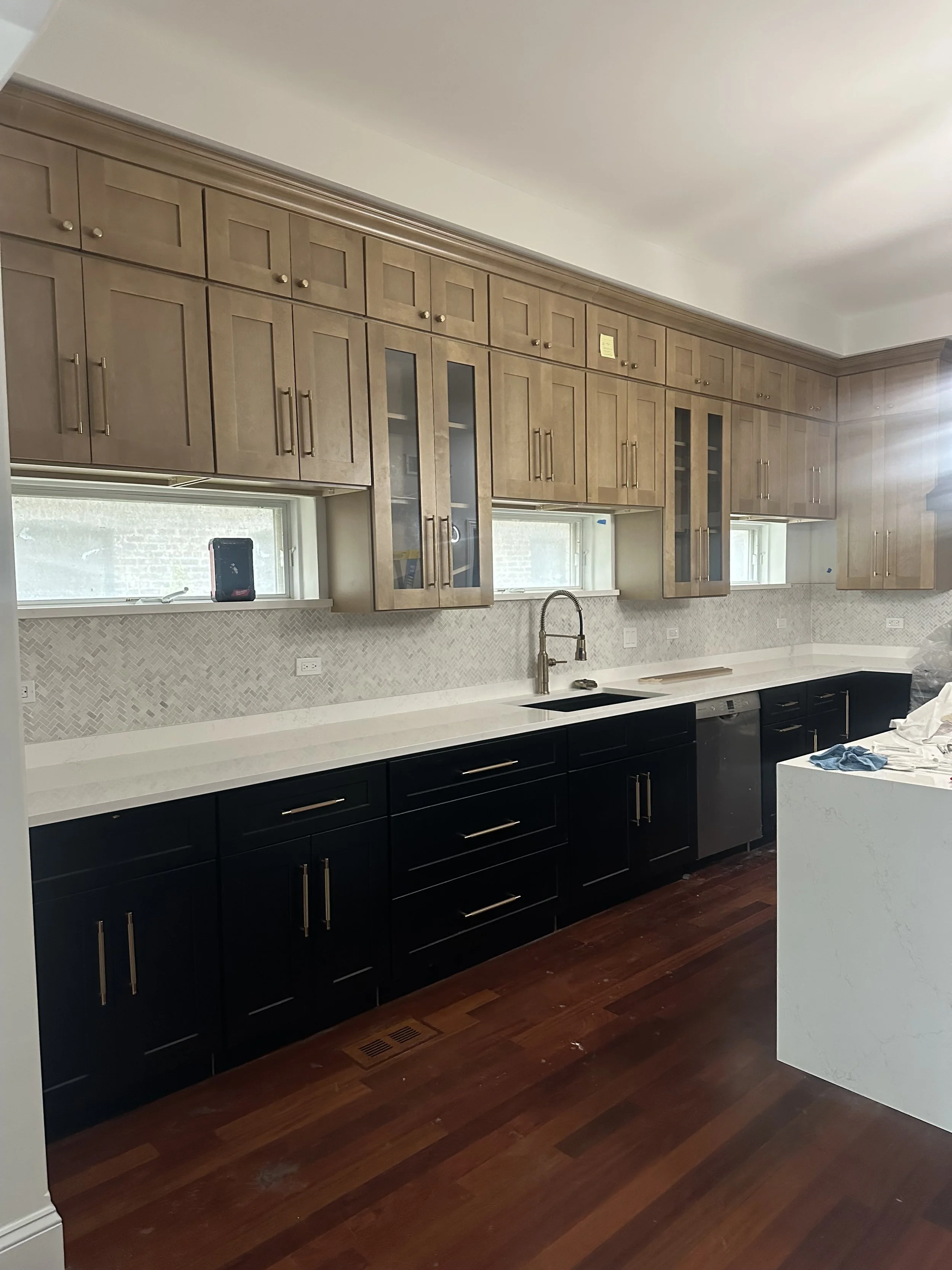 Kitchen with lower black cabinets, upper light wood cabinets, white countertop, backsplash, and hardwood floor.