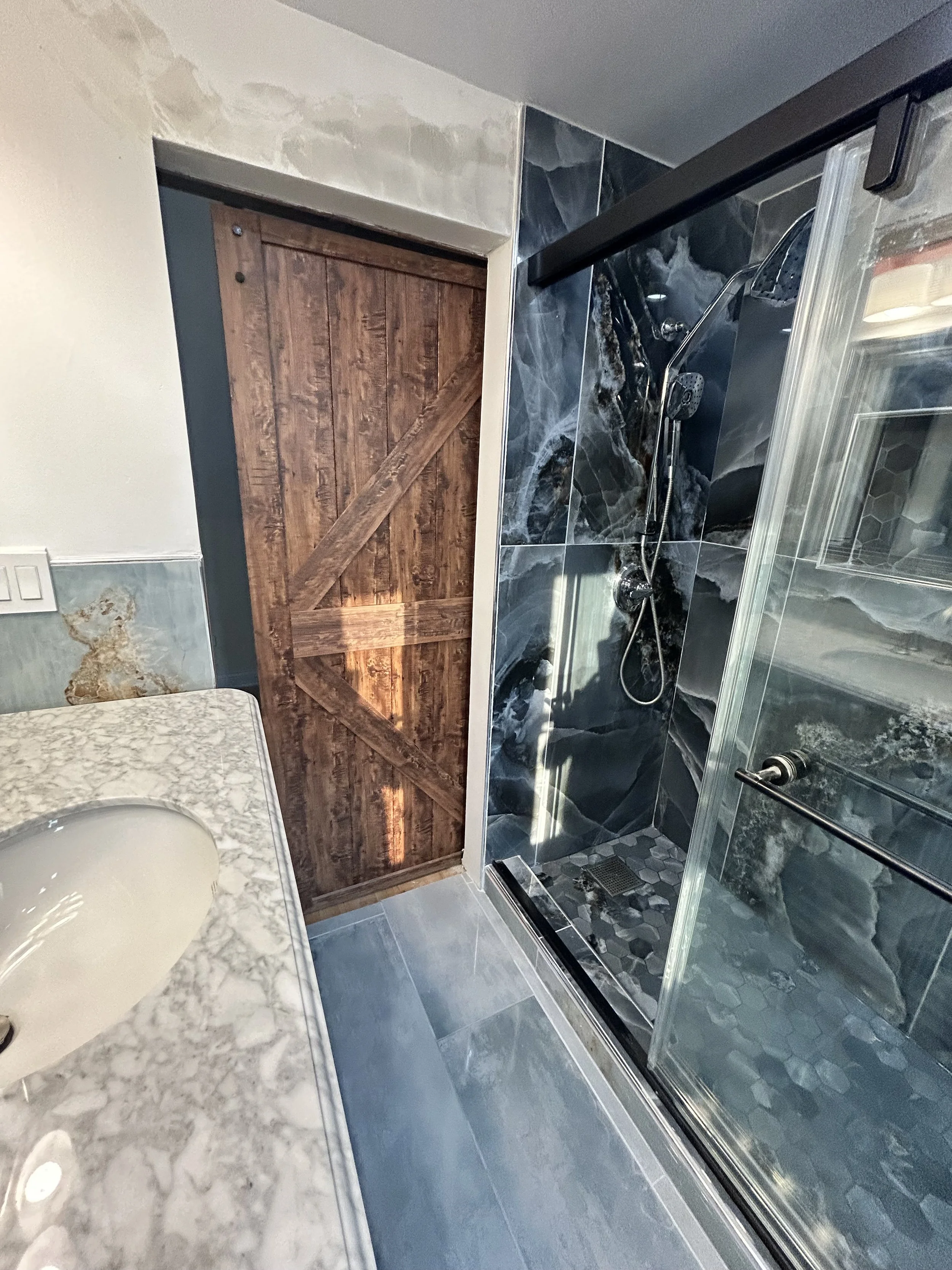 A bathroom with a marble countertop, a partial view of a sink, a wooden sliding door, and a shower stall with dark marble tiles, a glass door, and a handheld shower head.