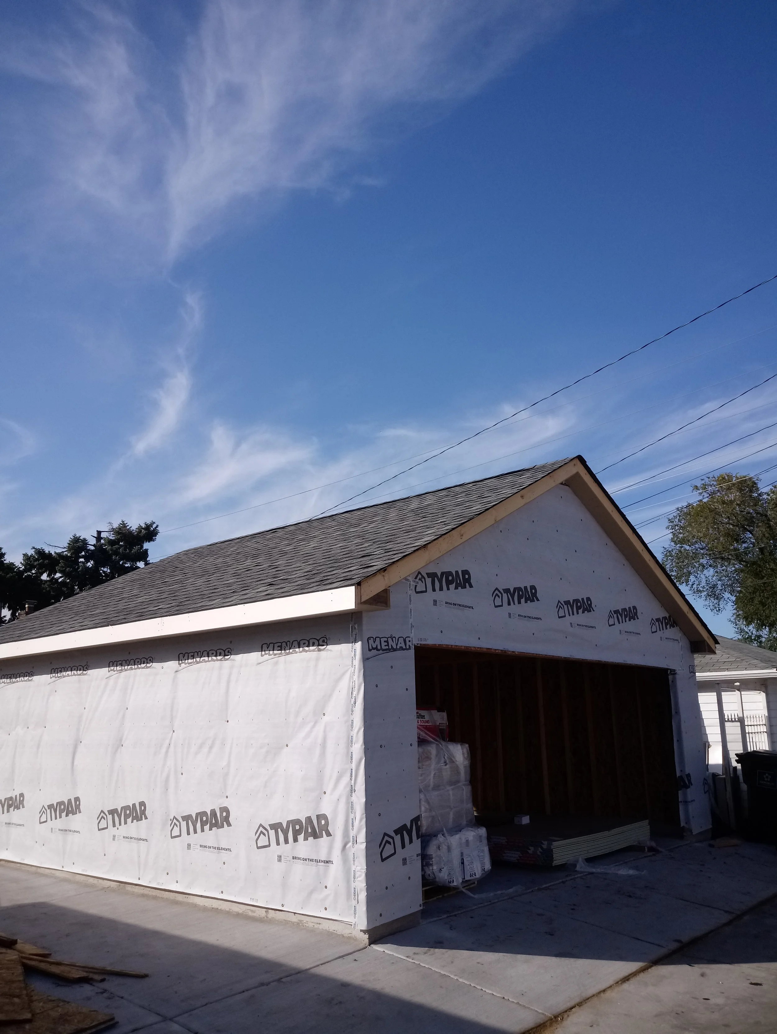 A house under construction with a partially built wall covered in Typar weather-resistant barrier, a roof with shingles, and an open garage area. The sky is clear with some wispy clouds, and there are power lines and trees in the background.