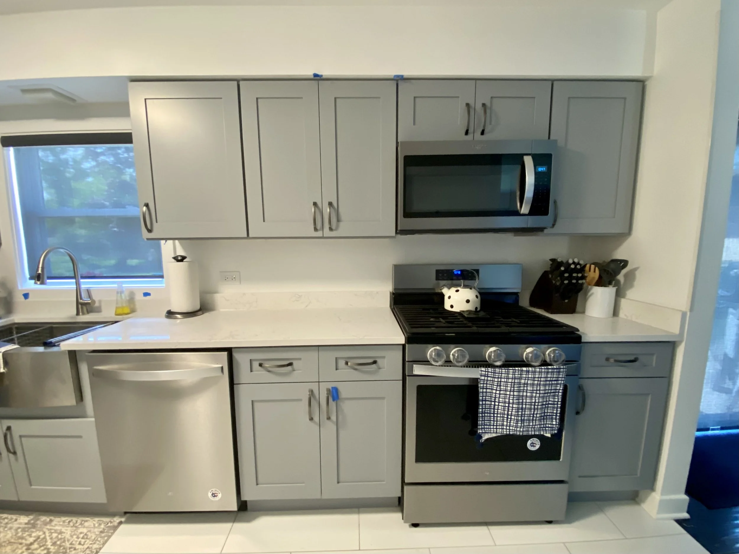 This image shows a modern kitchen with gray cabinets, a window with blinds, a stainless steel microwave, and a stove. There are kitchen utensils and a tea towel on the counter.