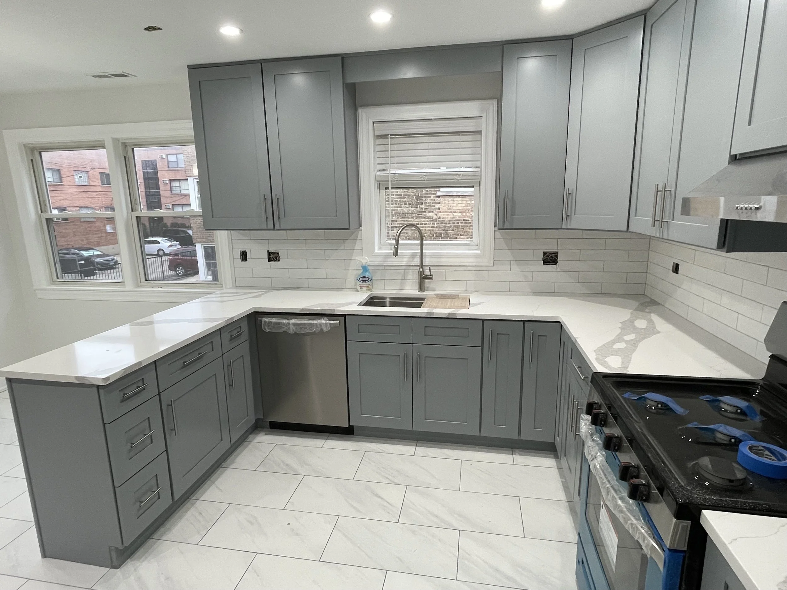Newly renovated kitchen with light gray cabinets, white marble countertops, and a white subway tile backsplash. Two large windows look out onto brick buildings and parked cars.
