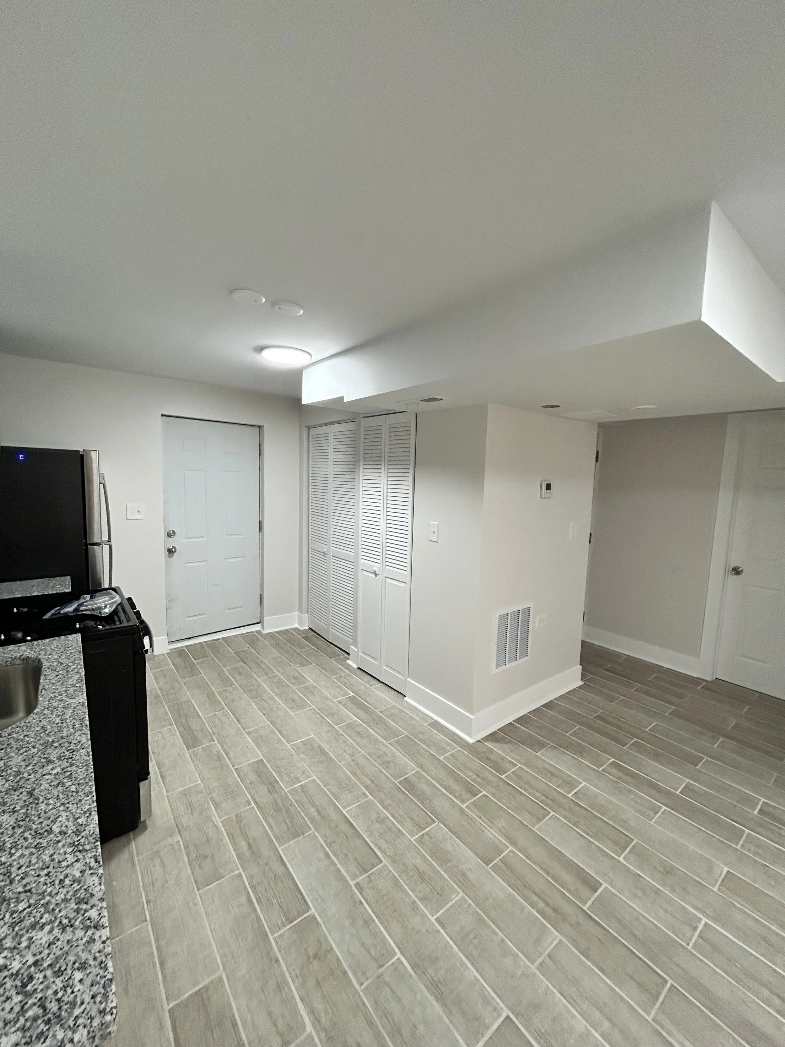 A clean, modern kitchen with gray tile flooring, white walls, a stainless steel refrigerator, and a white door. The ceiling has recessed lighting and a round ceiling light, with a vent near the wall.