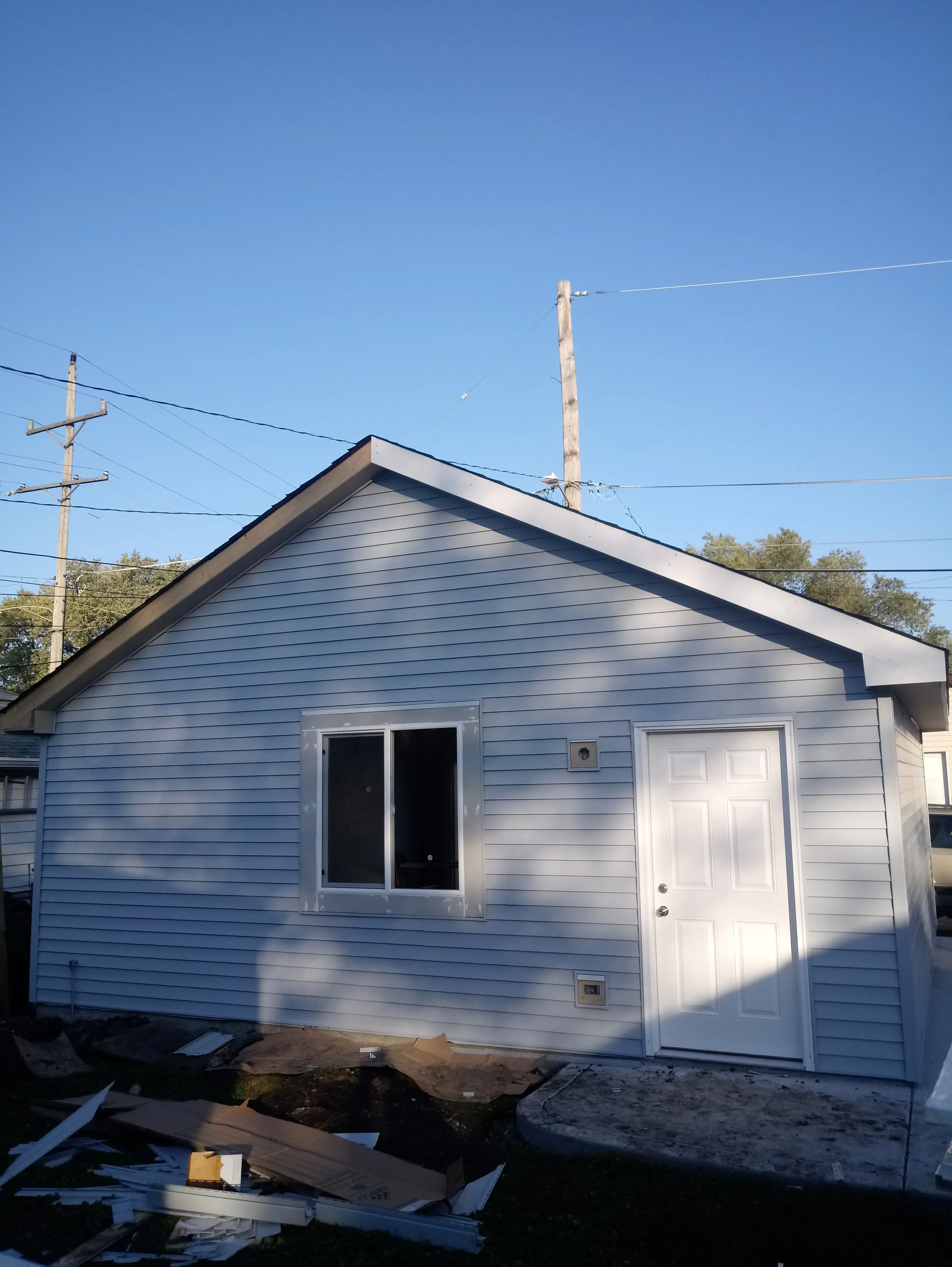 Back of a house with white vinyl siding, a window, a door, and debris including torn cardboard and white plastic on the ground, with power lines and utility poles against a clear blue sky.