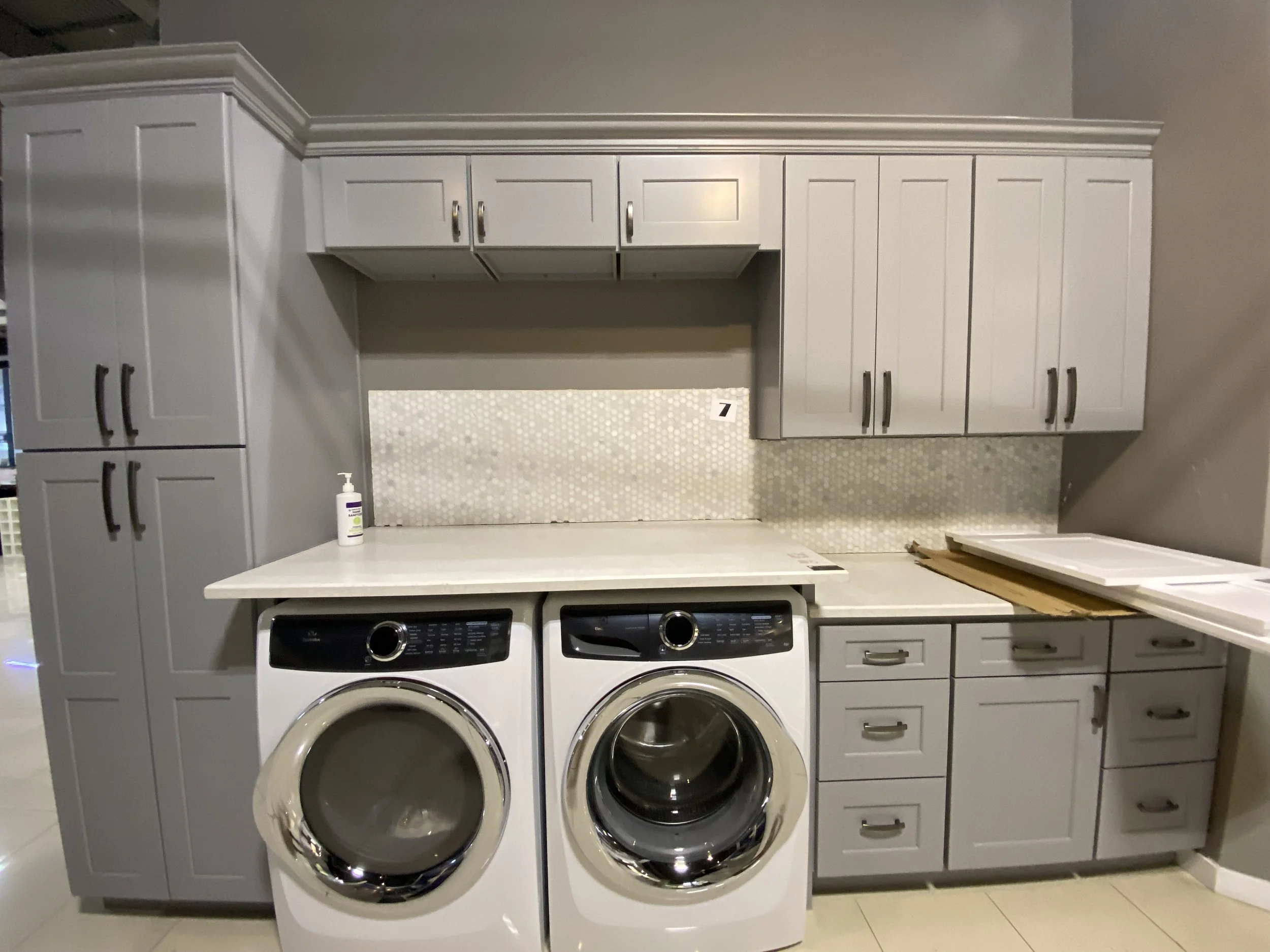 White laundry room with white cabinets above and drawers below, with two front-loading washers side by side, and a small countertop with a bottle of soap or sanitizer. There is a patterned backsplash and a piece of furniture or paper stacked on the r