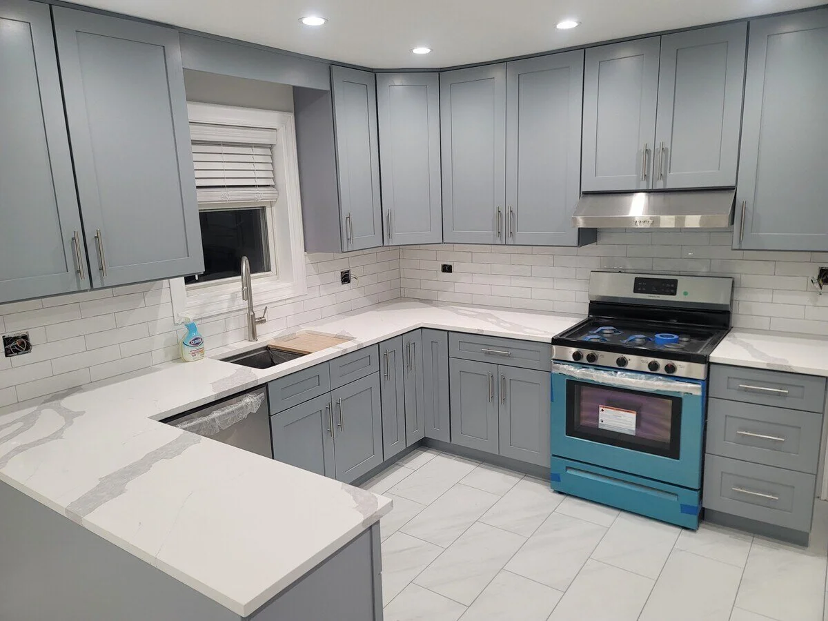 Modern kitchen with light gray cabinets, white marble countertops, a window with blinds, and a stainless steel oven with a range hood.