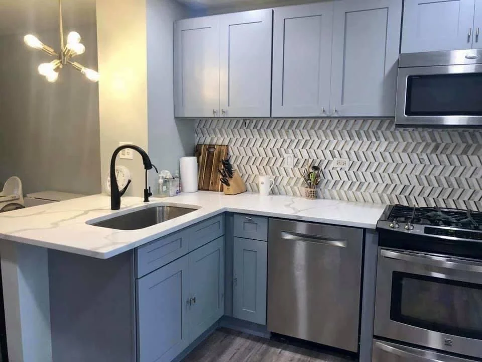 Modern kitchen with gray-blue cabinets, stainless steel appliances, and white marble countertop. Backslash with a herringbone pattern. Kitchen tools and paper towels on the counter.