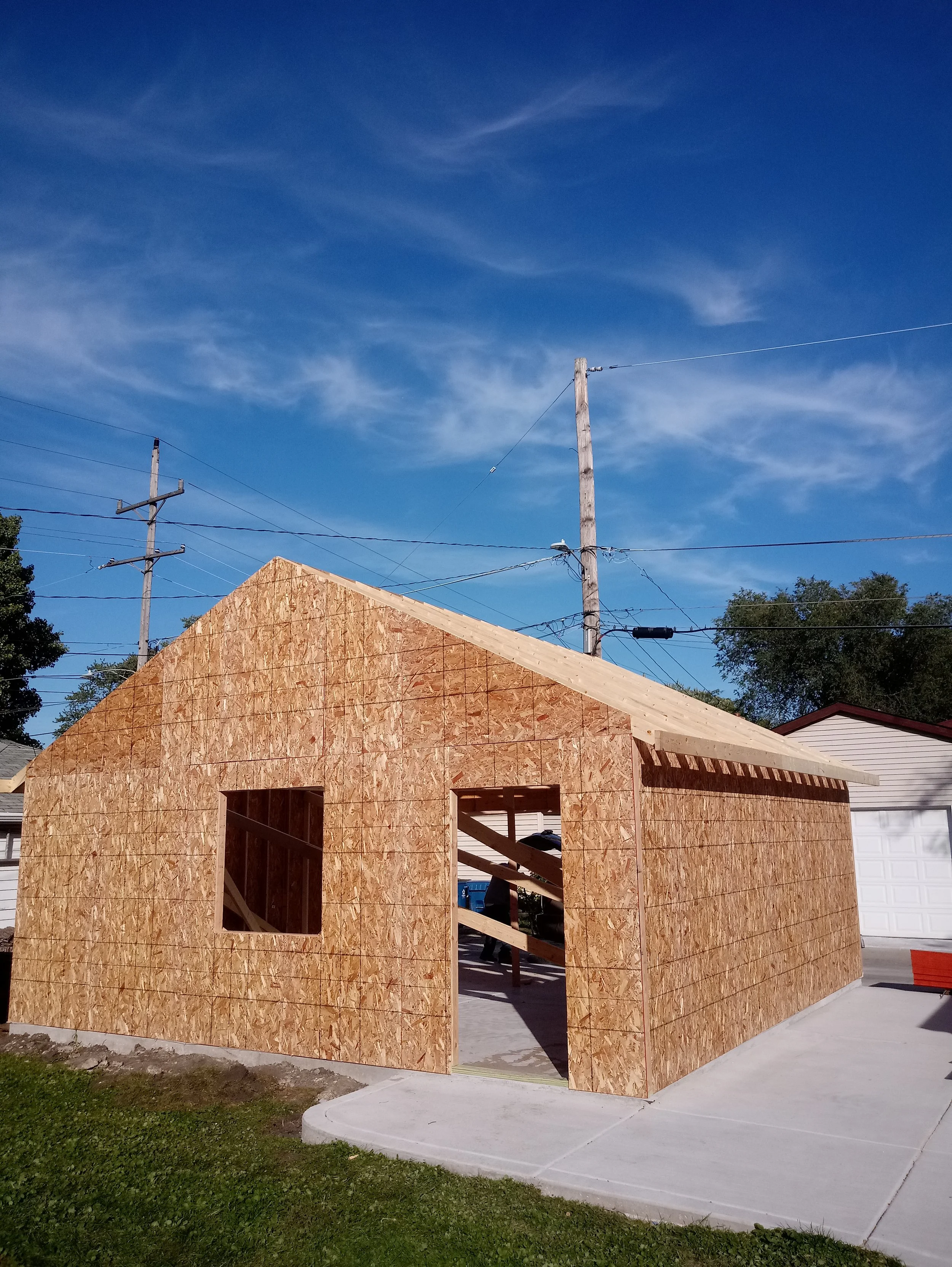 A house under construction with wooden walls, a concrete driveway, and utility poles in the background under a blue sky with scattered clouds.
