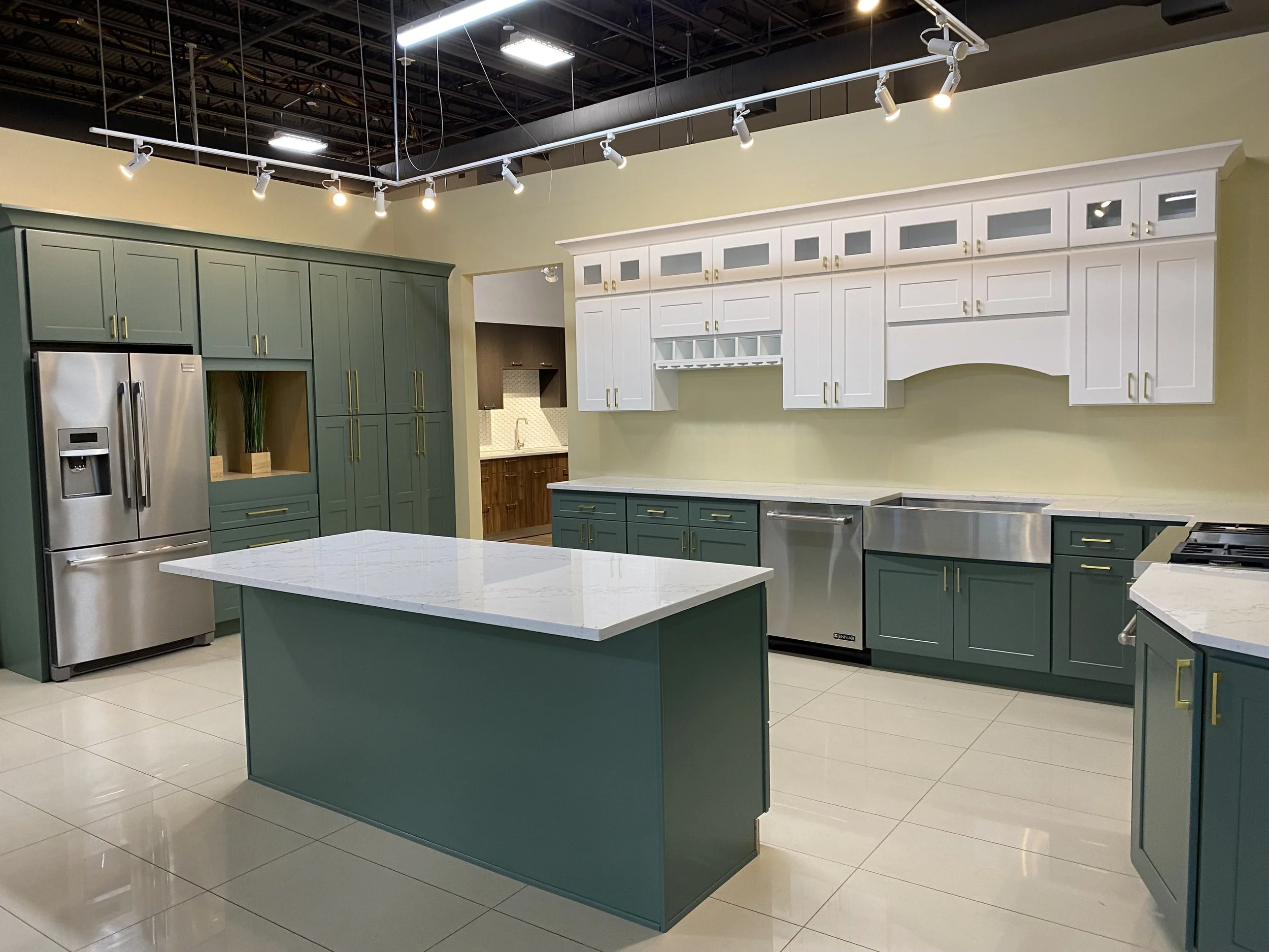 Showroom kitchen display with green cabinets, white upper cabinets, a kitchen island, stainless steel fridge, and hanging track lighting.