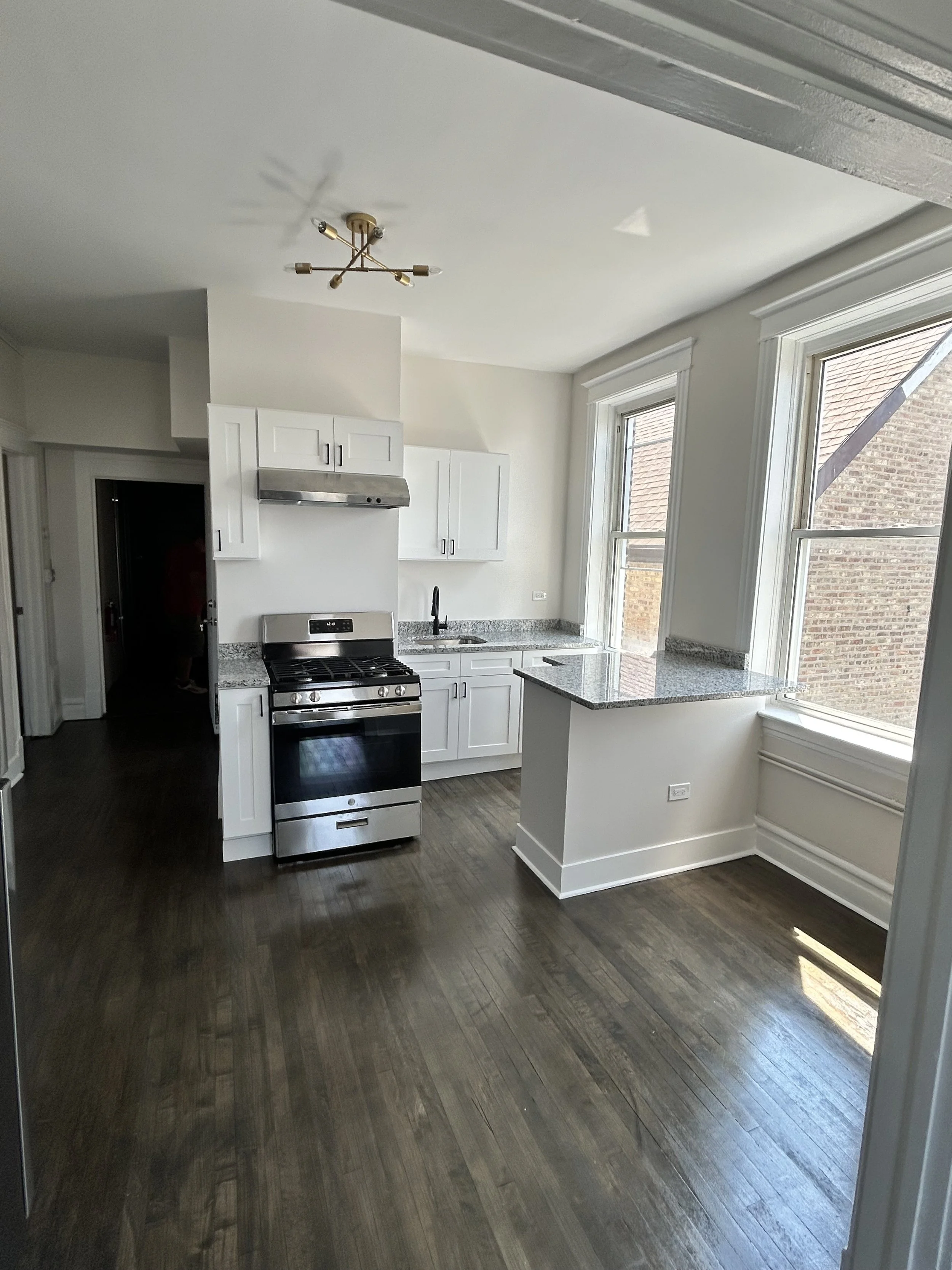 Empty kitchen with dark wood flooring, white cabinets, granite countertops, stainless steel stove, range hood, black faucet, and large windows.
