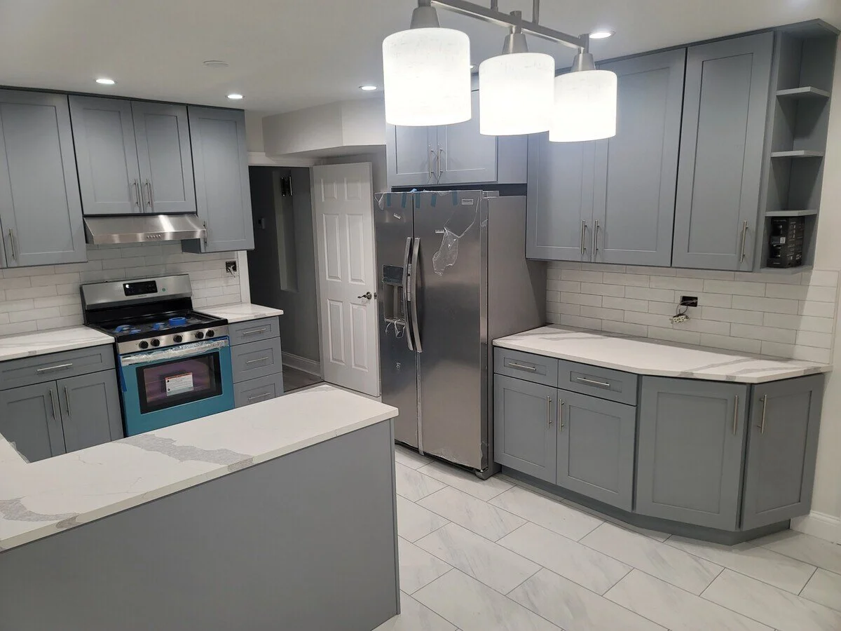 Modern kitchen with gray cabinets, white countertops, and stainless steel appliances. Overhead lighting with three white pendant lights.