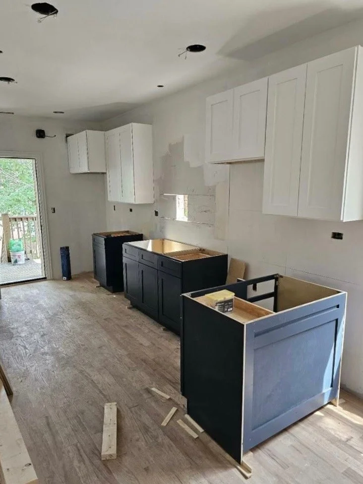 Kitchen under renovation with white upper cabinets, navy lower cabinets, and a wooden floor. Some cabinets are missing doors, and construction materials are on the floor.