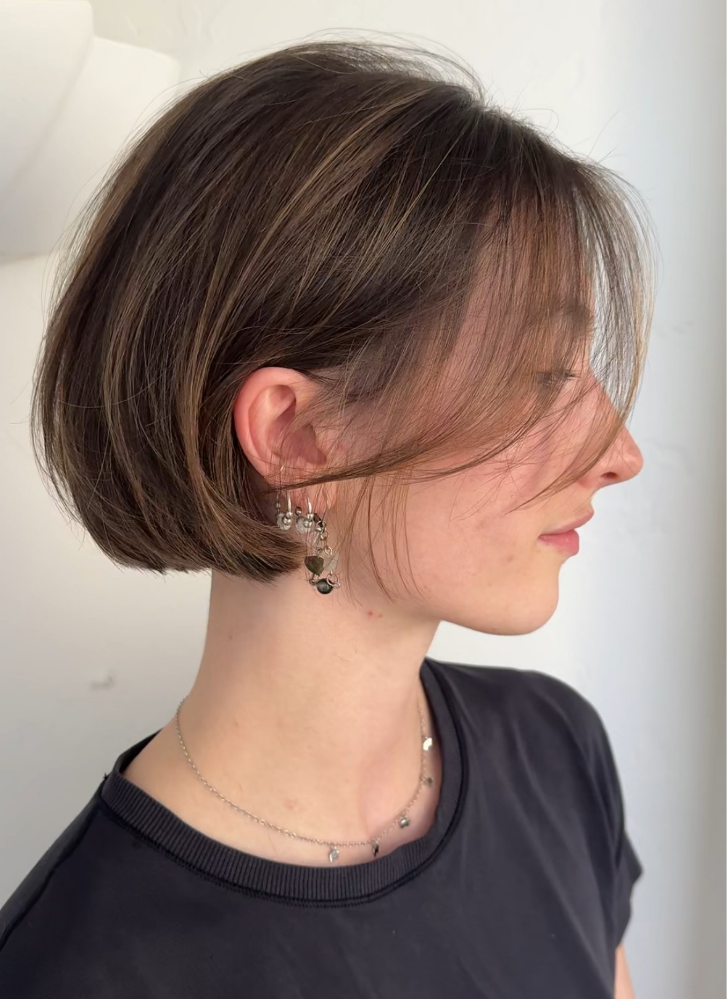 Side profile of a woman with a short, layered brown hairstyle, wearing a black shirt and jewelry, against a plain light background.