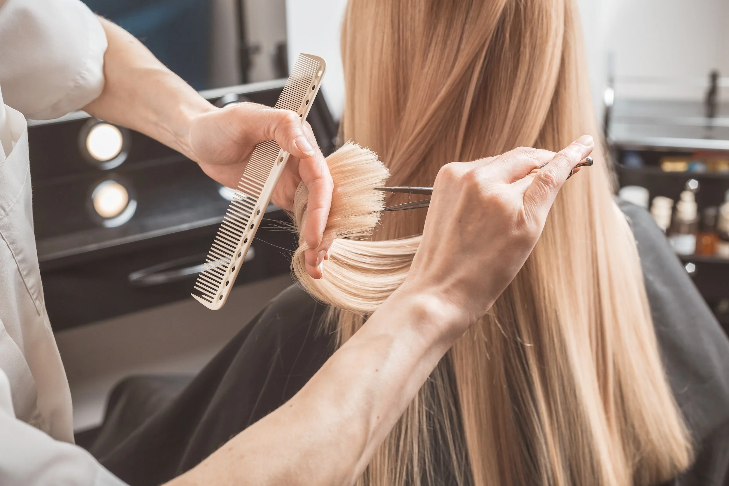 A hairstylist holding a comb and scissors while cutting a woman's long, blonde hair in a salon.