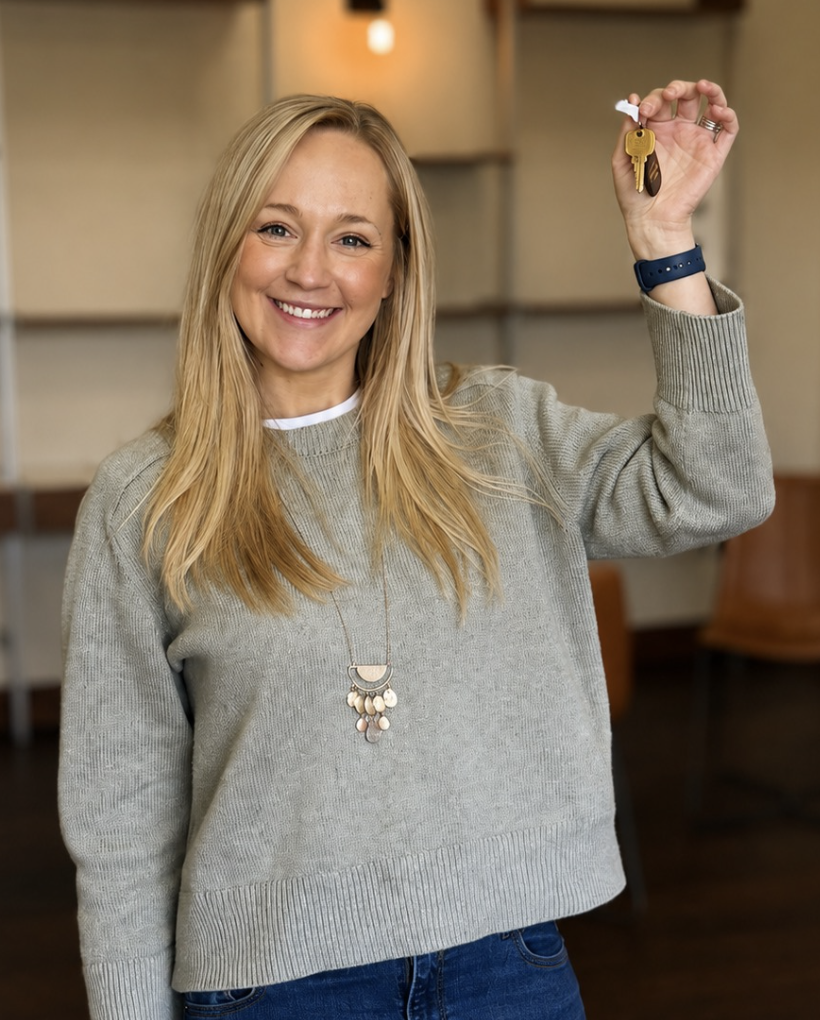 A smiling woman with blonde hair holding keys with a keychain in a room with wooden furniture and shelves.