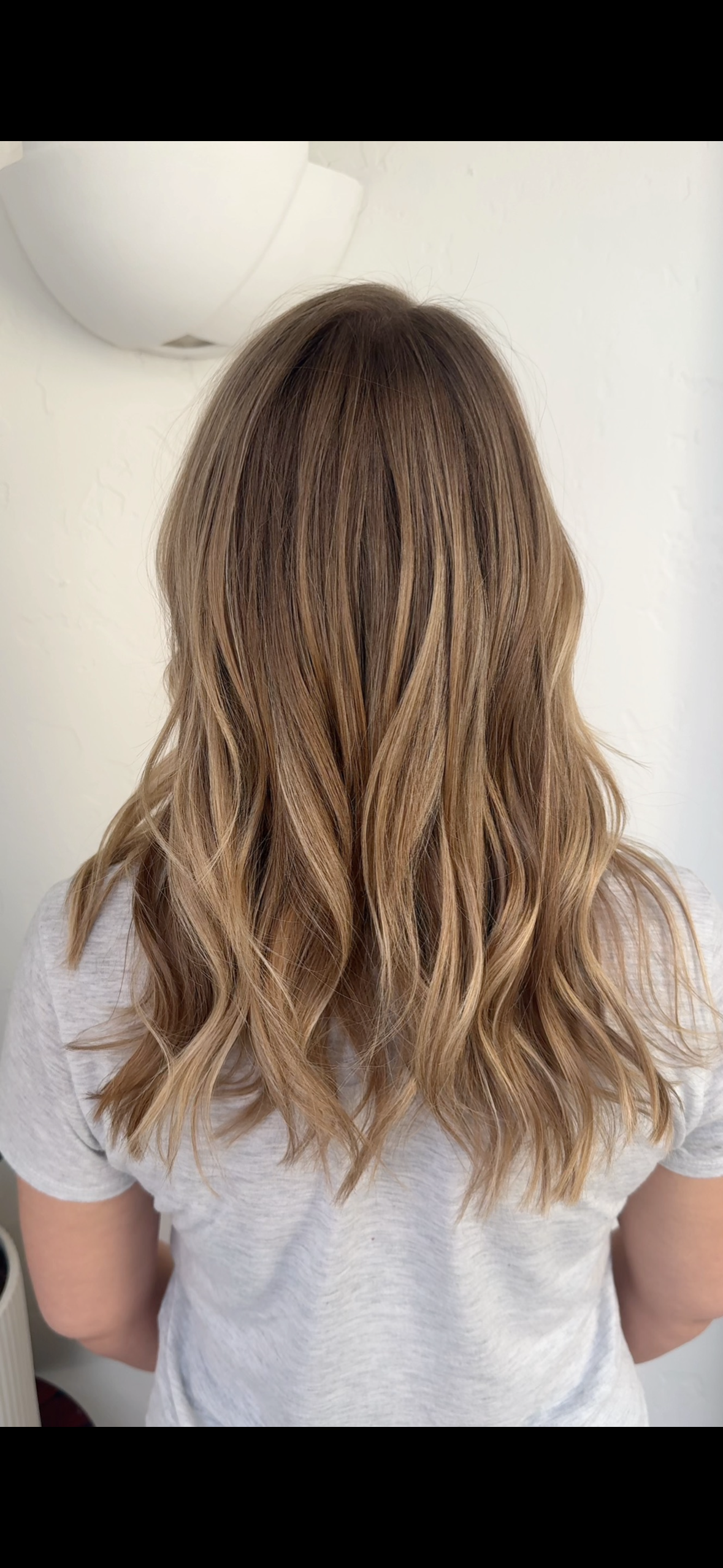 Back view of a woman with shoulder-length, wavy blonde hair standing against a plain white wall after balayage treatment