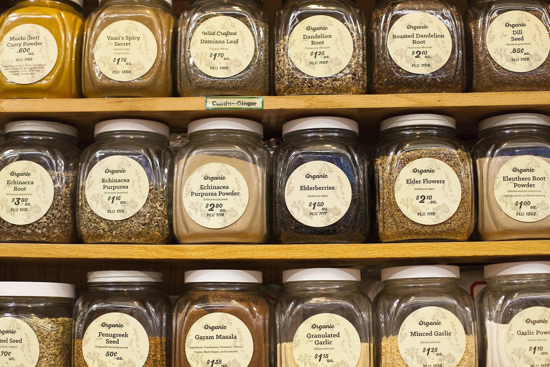 Several jars of organic herbs and spices on wooden shelves at a store. The jars are labeled with names such as Yukon Spicy Secret, Wild Crafted Damiana Leaf, Dandelion Root, Roasted Dandelion Root, Dill Seed, Echinacea Root, Echinacea Purpurea, Elderberries, Elderflower, and Garam Masala, with prices and product details.