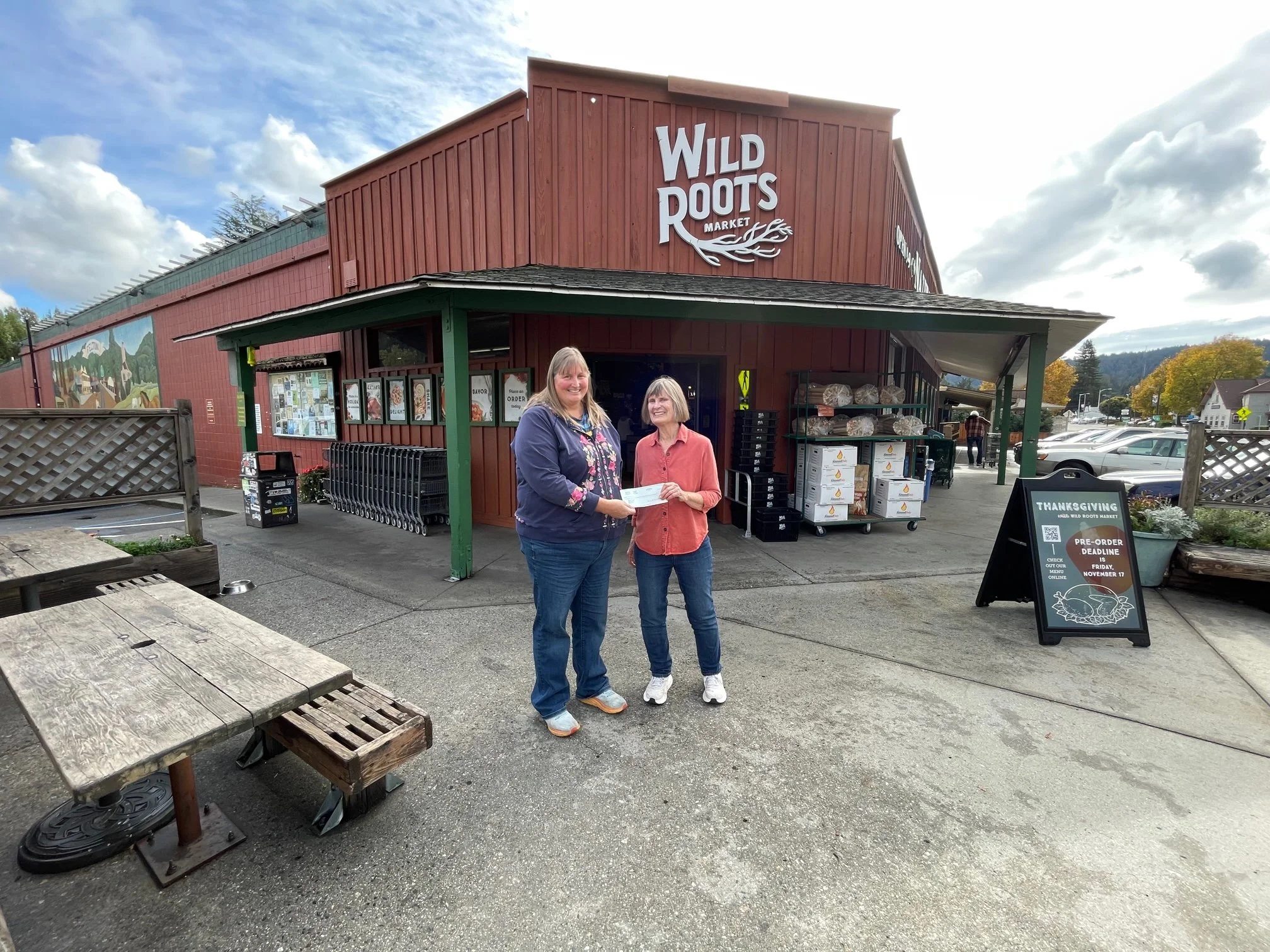 Two women stand in front of Wild Roots Market, a building with a red wooden exterior, while holding a check. The woman on the left has long blond hair and is wearing a navy jacket with floral details, and the woman on the right has a bob haircut and is dressed in a coral-colored shirt. There are store notices and shelves with goods outside the store, and parked cars in the background.