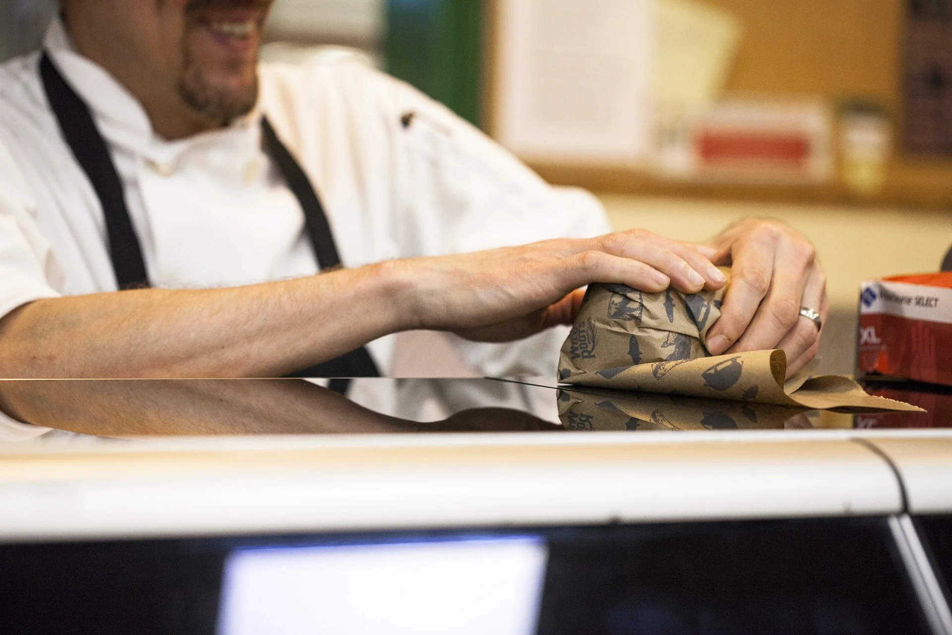 A person in a white chef uniform wrapping a sandwich in decorative paper on a kitchen counter.