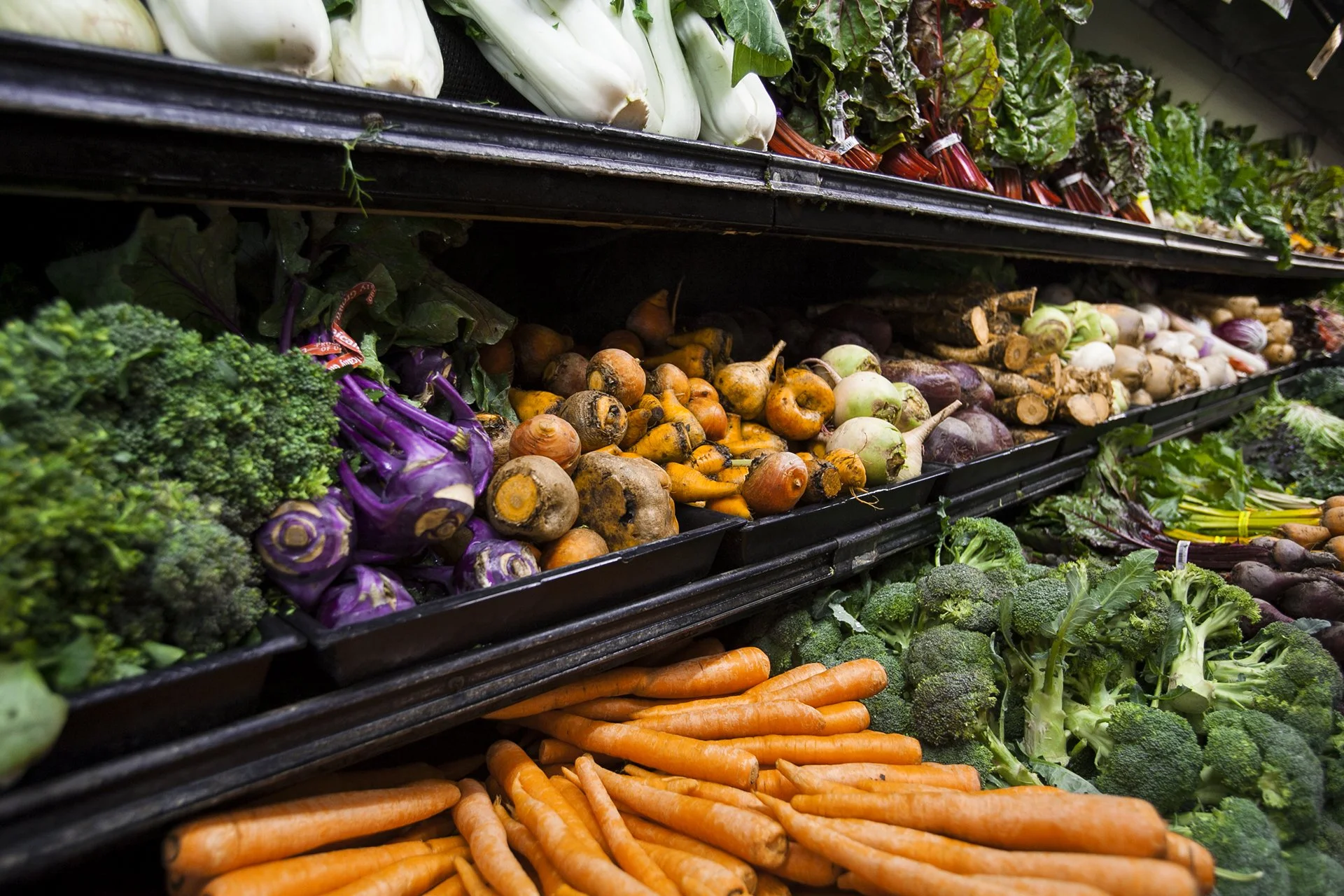 Display of various fresh vegetables at a grocery store, including carrots, broccoli, onions, cauliflower, purple eggplants, and leafy greens, arranged on black trays.