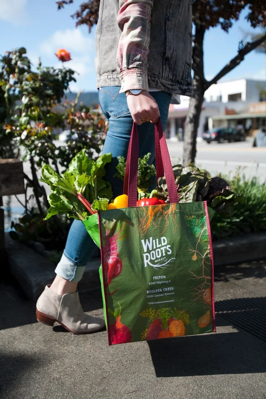 A person holding a reusable shopping bag filled with fresh vegetables outdoors on a sunny day. The bag has the logo "Wild Roots Market" and contains leafy greens and colorful peppers. The person is wearing jeans, a denim vest, and ankle boots.