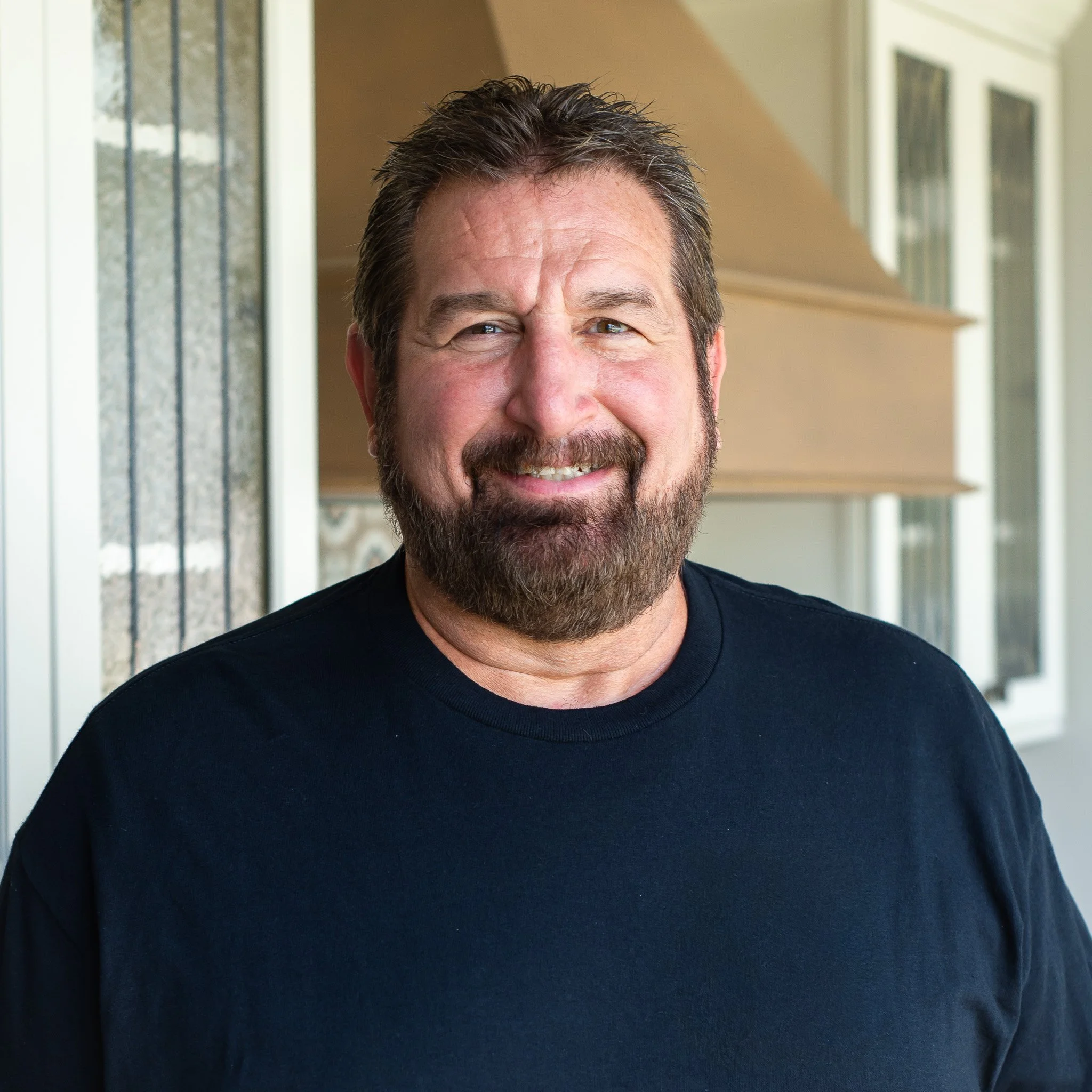 A smiling man with dark, graying hair and a beard, wearing a black shirt, standing outdoors with windows and walls in the background.