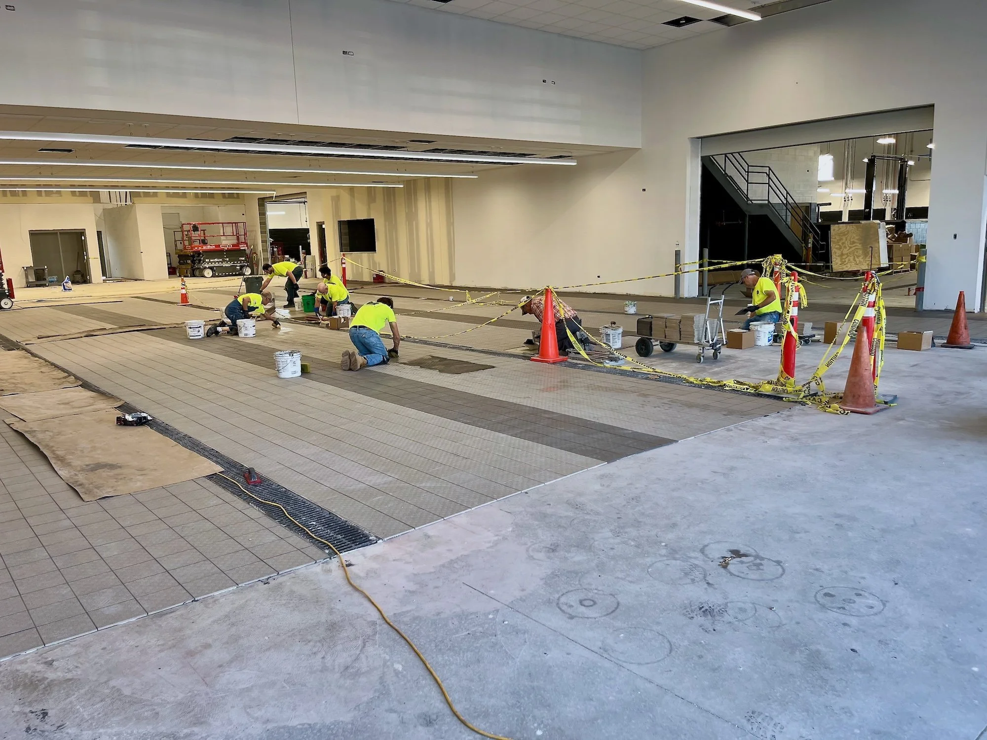 Construction workers installing large tiles on a floor inside a building, with caution tape and orange cones surrounding the work area.