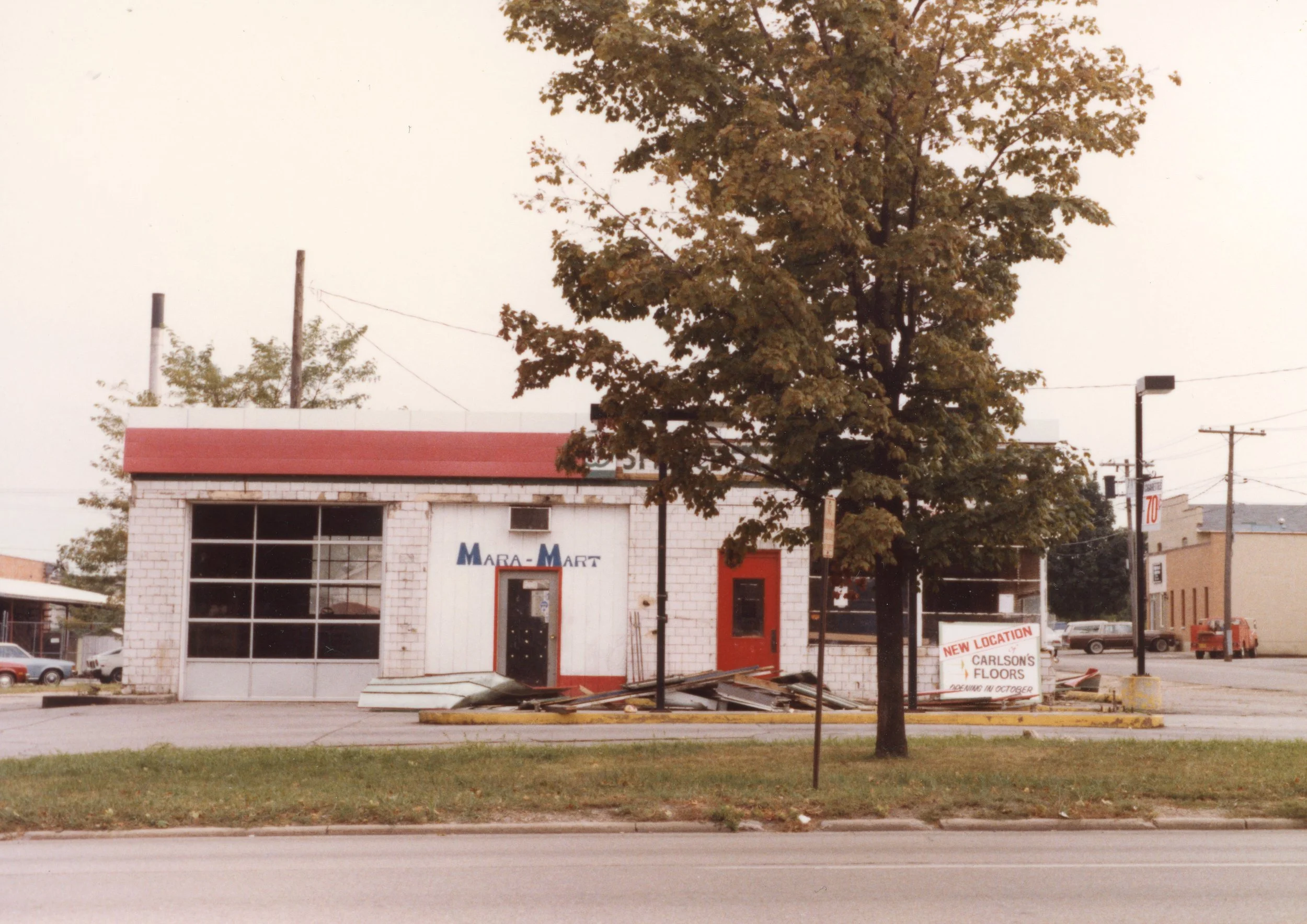 A small building with a red roof and white brick walls, with a sign indicating a new location for Carlson's Floors under construction, and a large tree in front.