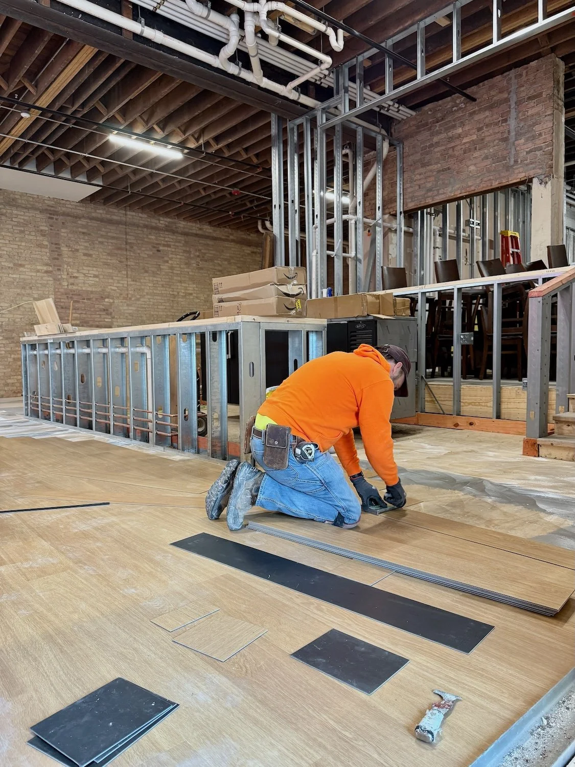 A construction worker kneels on the floor installing laminate flooring in an unfinished room with exposed brick walls, metal framing, and construction materials around.