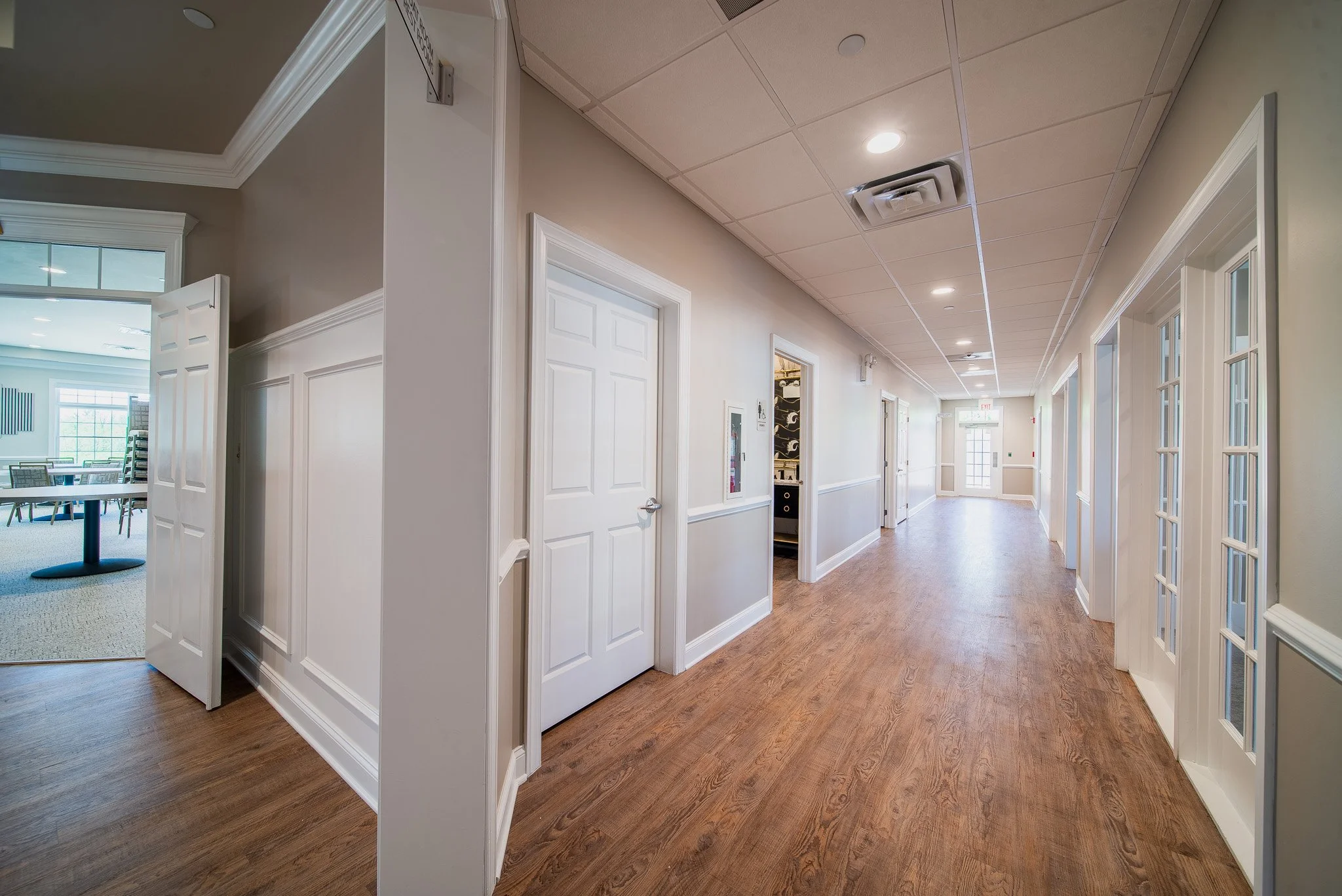Hallway in a building with white walls, wooden flooring, ceiling lights, and multiple doors along the corridor.