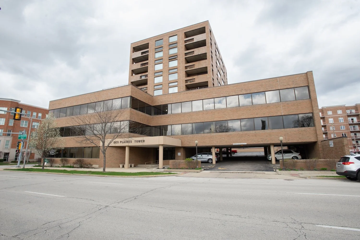 A multi-story brick building named Des Plaines Tower, with parking on the ground level and trees in front, under a cloudy sky.
