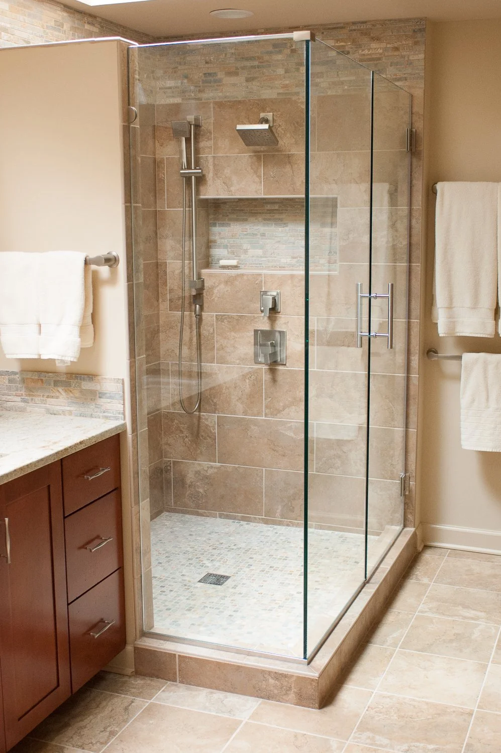 Enclosed walk-in shower with beige tile walls, a built-in tile shelf, a rainfall showerhead, a handheld showerhead, and a glass door handle, located next to wooden cabinetry and towel racks with white towels.