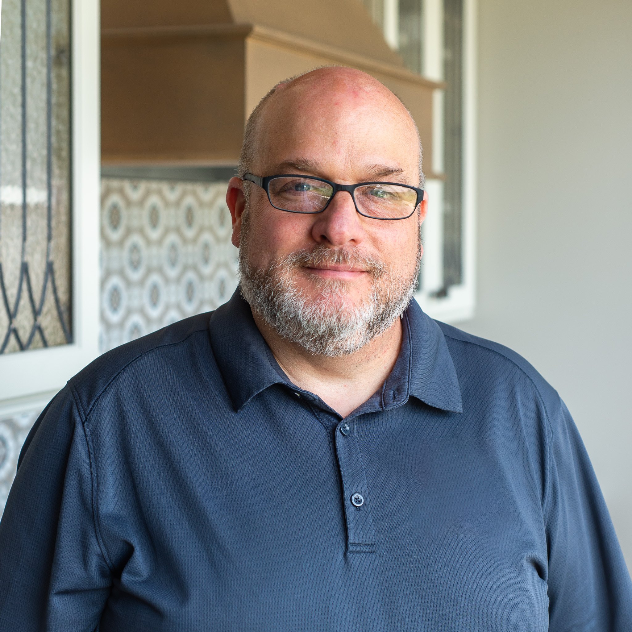 Close-up of a smiling man with glasses and a beard, wearing a dark blue shirt, standing in front of a window with patterned curtains and a beige wall.