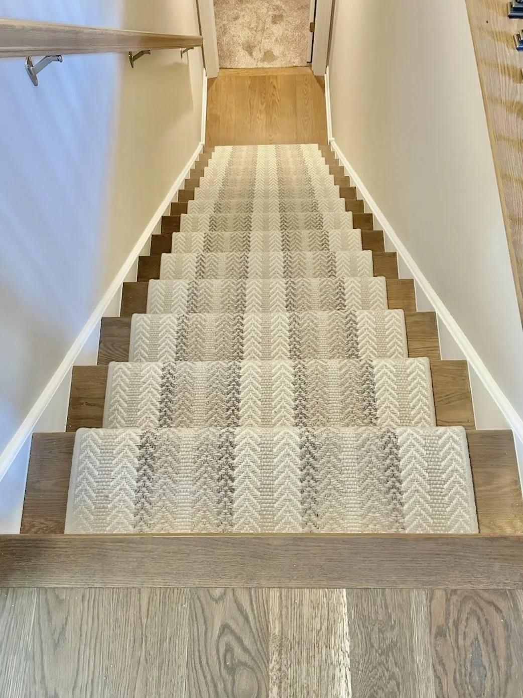 Top view of carpeted staircase with a beige and brown geometric pattern, bordered by wooden steps, leading down to a wooden floor and a doorway.