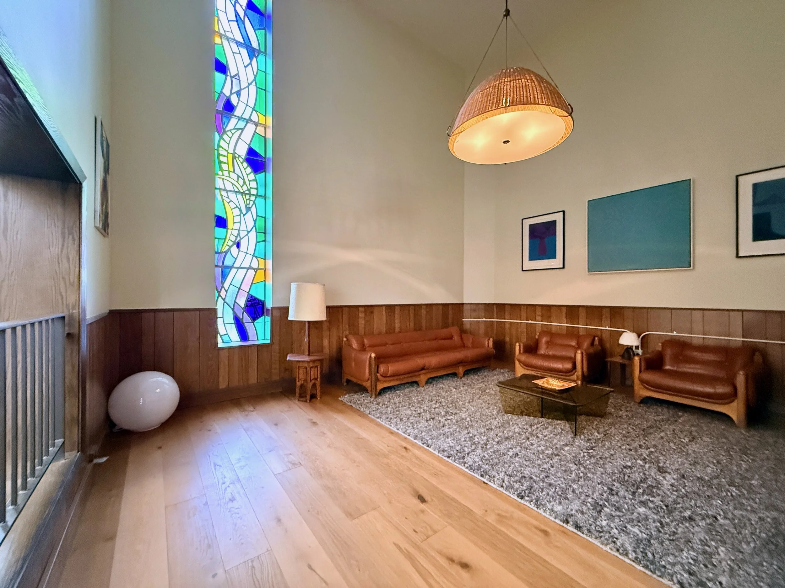 A cozy living room with wooden floors, leather sofas, a large grey rug, wall art, a tall colorful stained glass window, a hanging lampshade, a floor lamp, and spherical decorative objects.