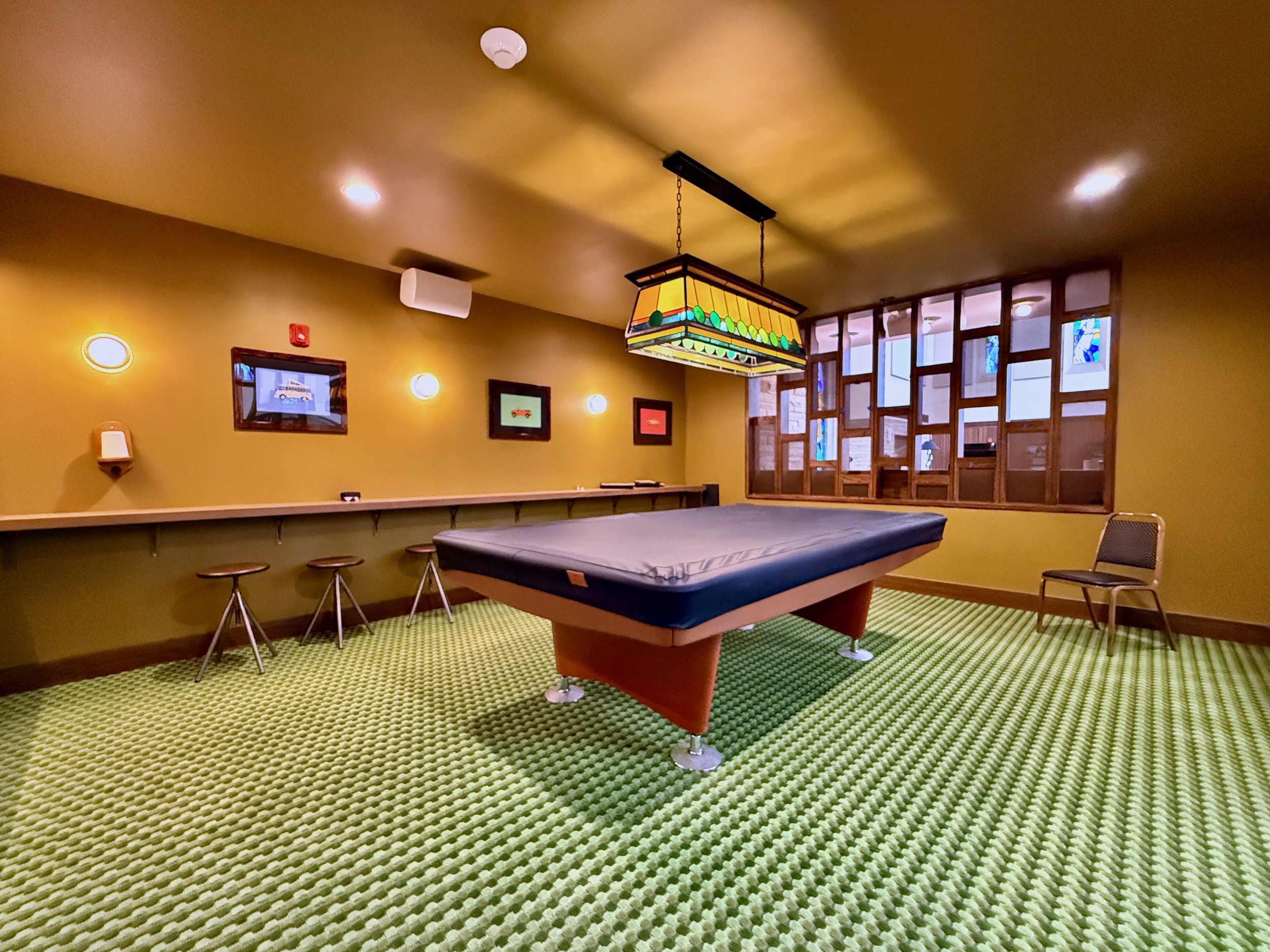 A room with a green textured carpet, a pool table, a stained glass hanging light, and several framed pictures on a yellow wall.