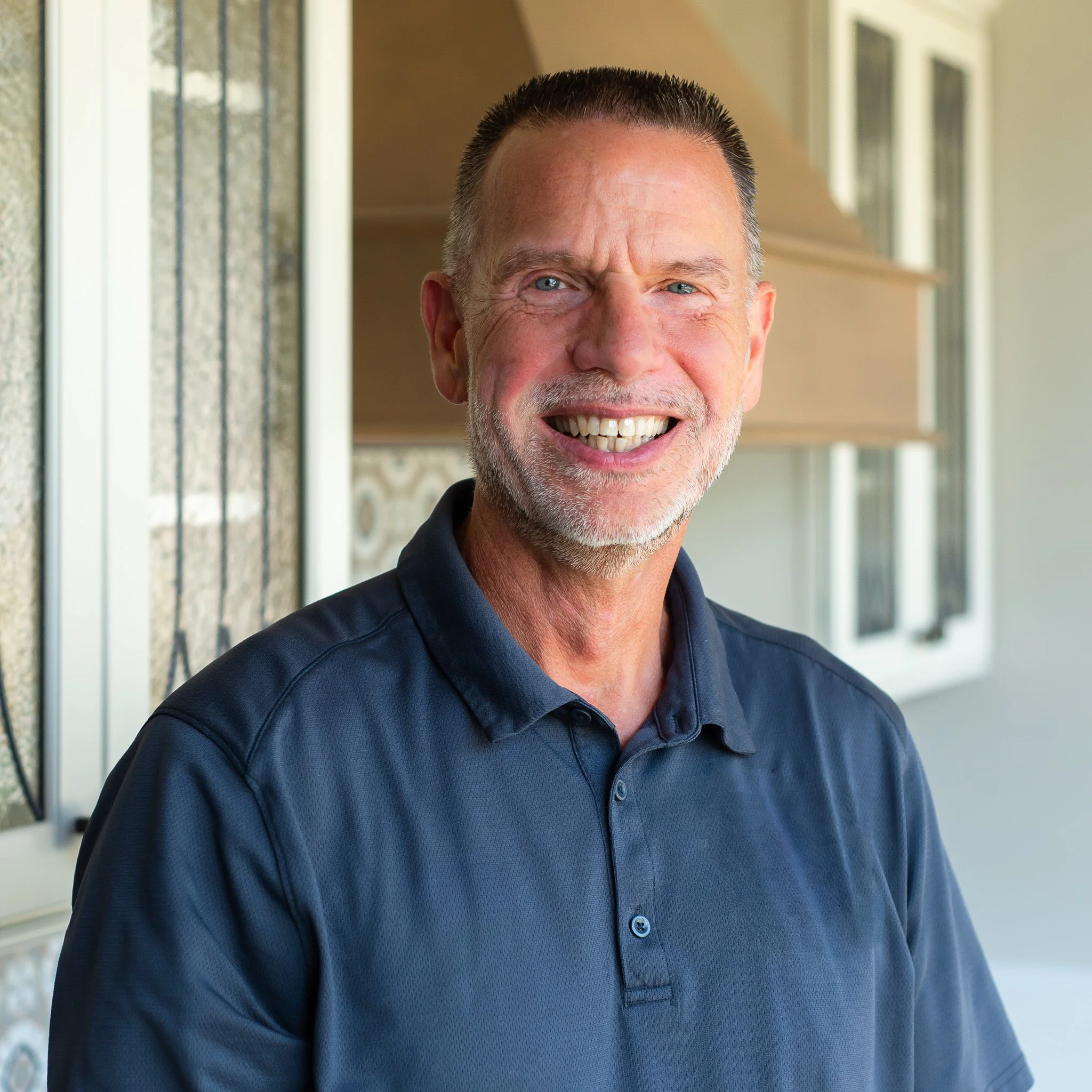 Close-up of smiling middle-aged man with short dark hair, blue eyes, wearing a navy blue collared shirt, outdoors near a house with windows and a porch.