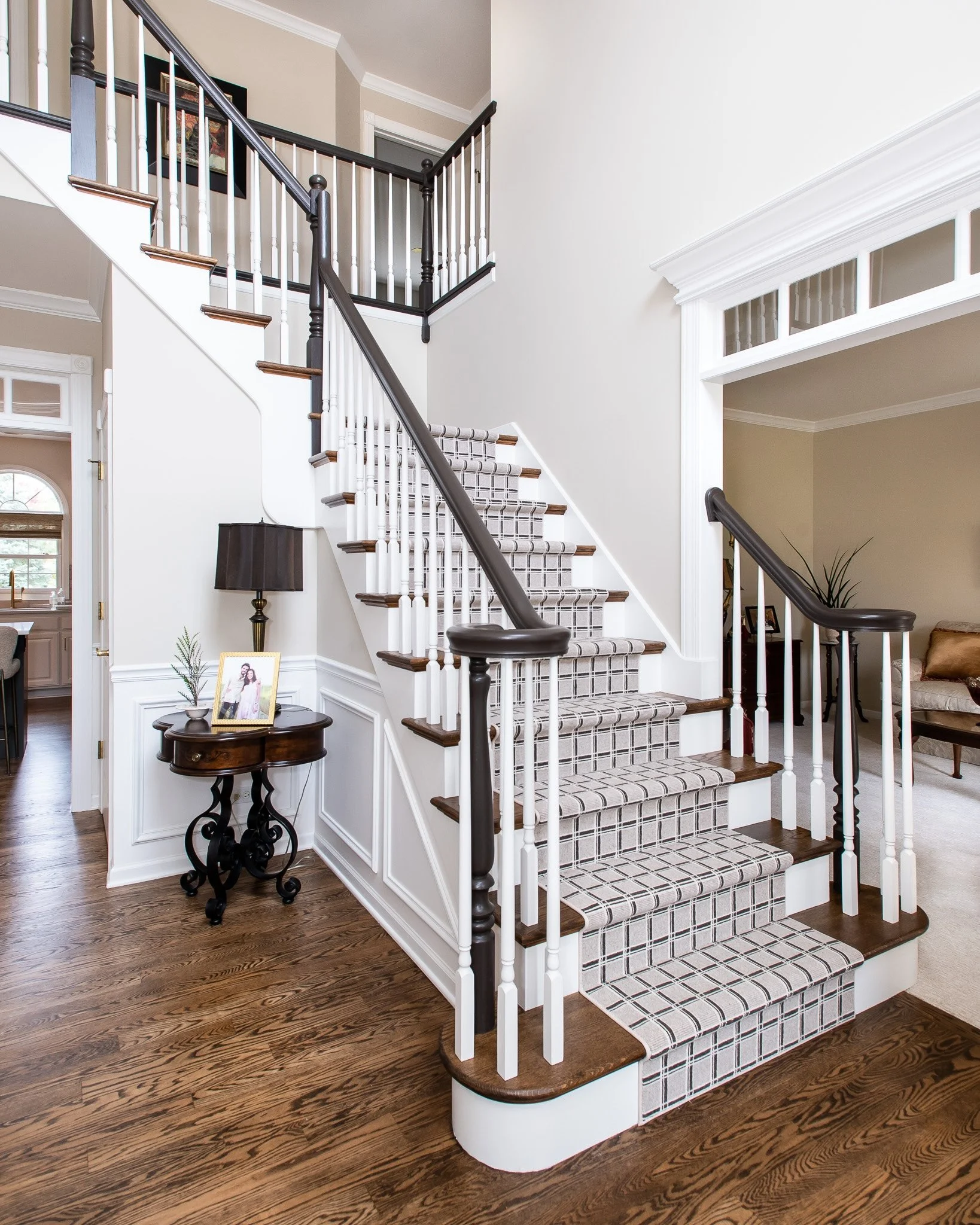 Interior view of a staircase with dark wood steps, white risers, black and white patterned runner, black handrails, white spindles, and decorative newel posts, leading to an upstairs balcony with white railing. A side table with a framed photo, lamp,